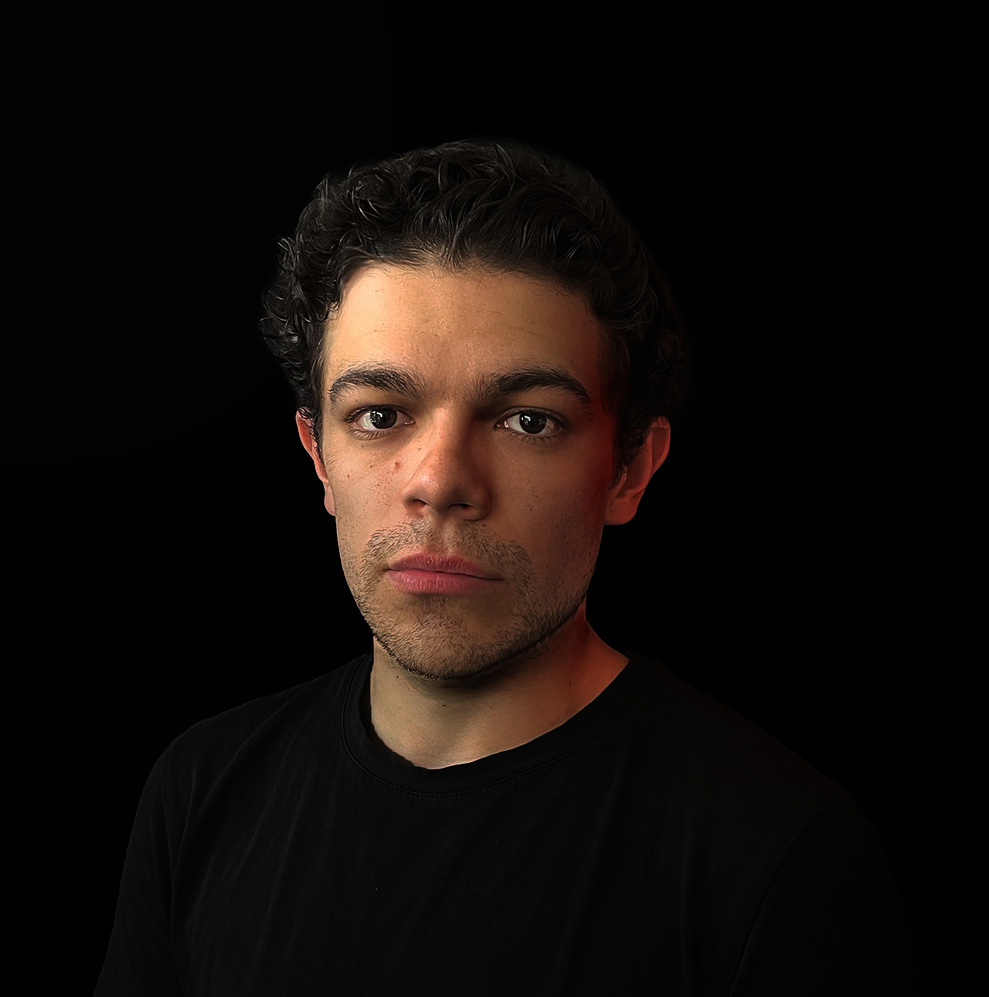 Portrait of a young man with curly dark hair and slight facial stubble wearing a black shirt against a black background.