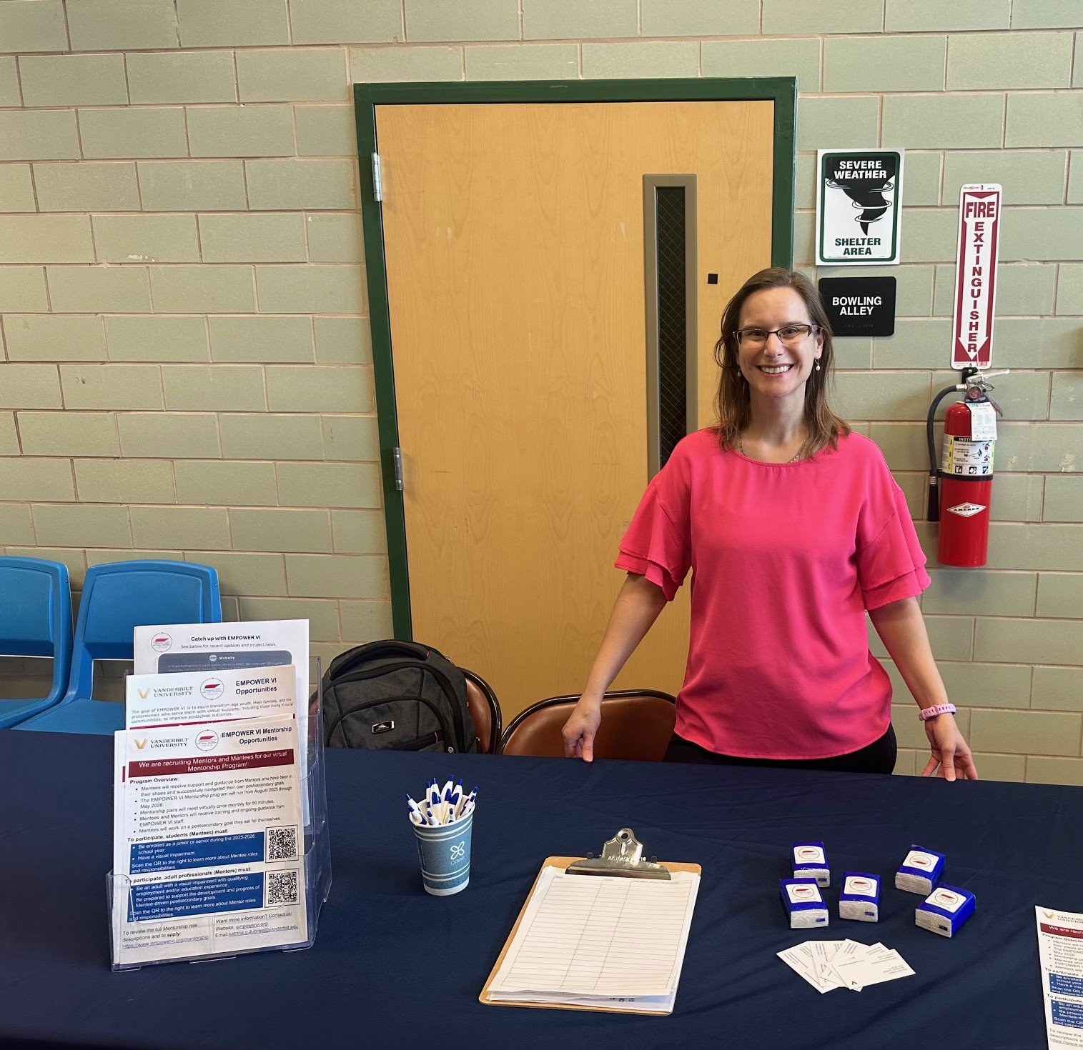 Katrina Dubree stands behind a table topped with flyers, pens, business cards, tissues, and an email sign-up sheet. Each item promotes the EMPOWER V I program. 