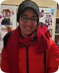 Sydney is a dark haired woman with brown skin wearing glasses and a red coat.