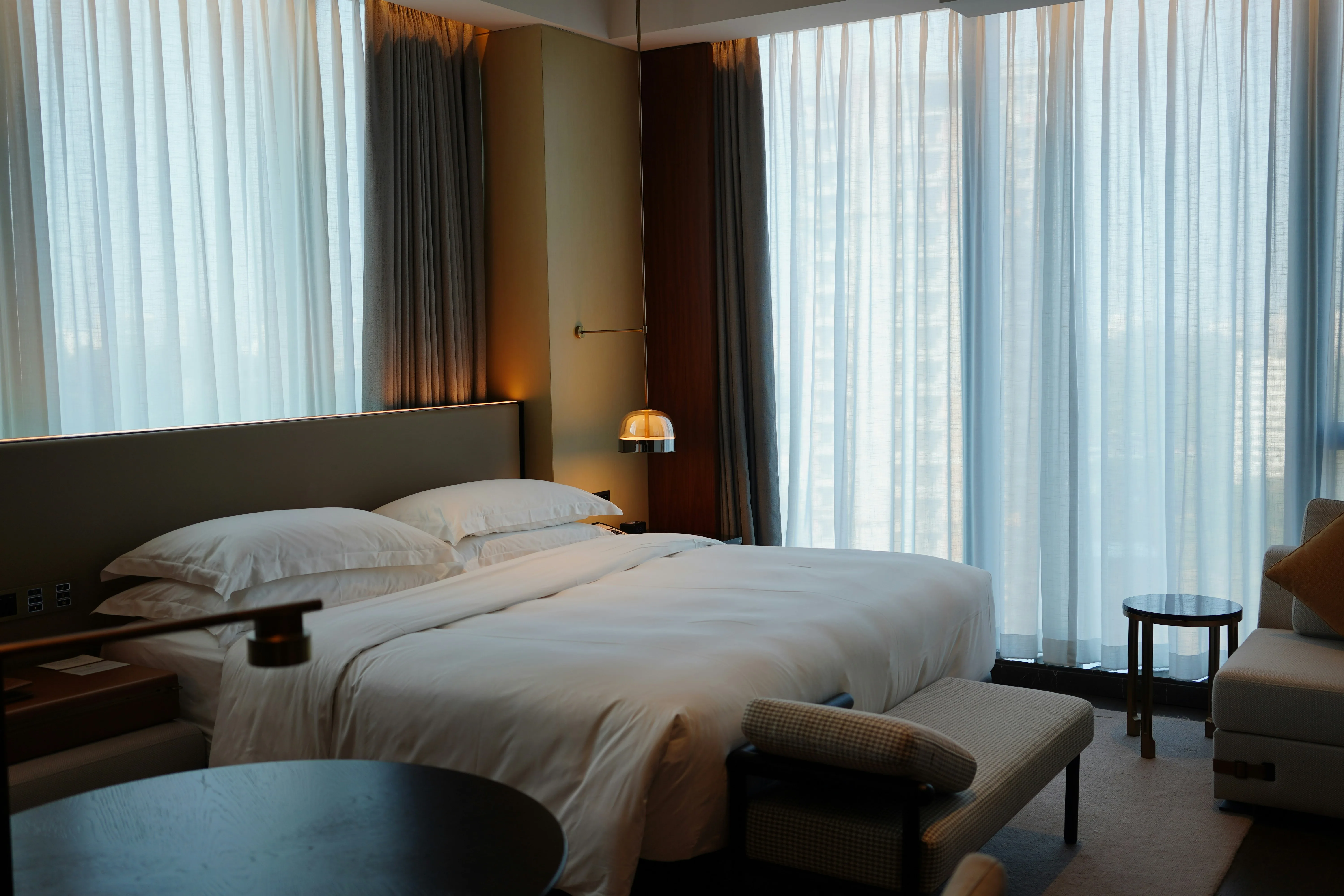 Camera de hotel moderna cu pat dublu, lenjerie alba si draperii care acopera ferestre mari. Foto: Travis Chen / Unsplash