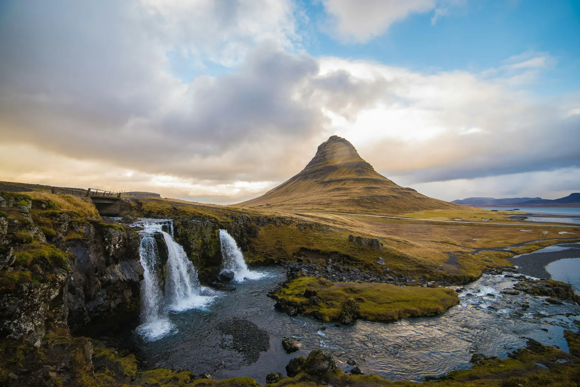 Muntele Kirkjufell si cascada Kirkjufellsfoss in Islanda, cu pajisti verzi si cer dramatic cu nori. Foto: Chris Ried / Unsplash