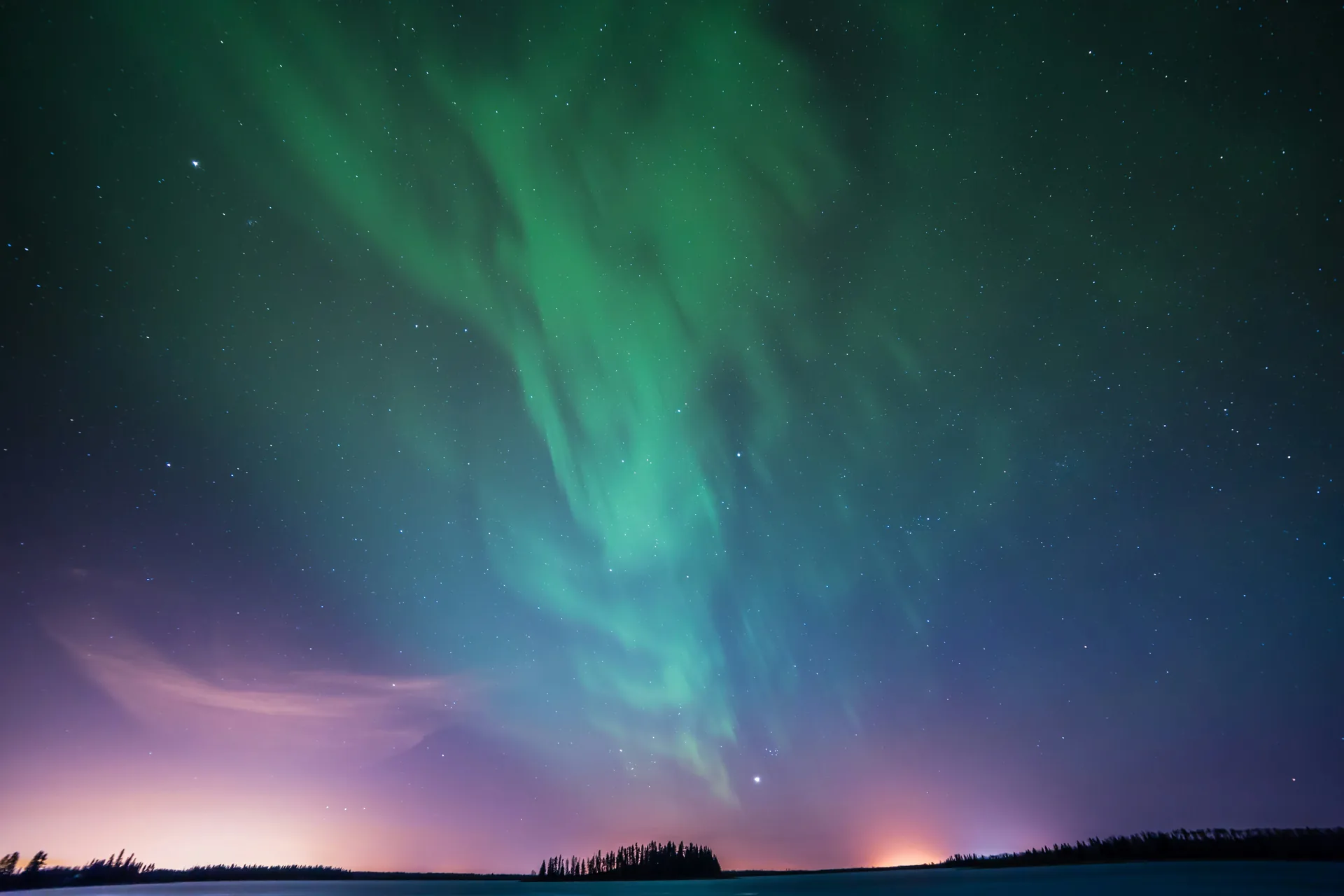 Aurora boreala verde si violet pe cerul instelat, cu silueta padurii boreale la orizont si lumini portocalii.
