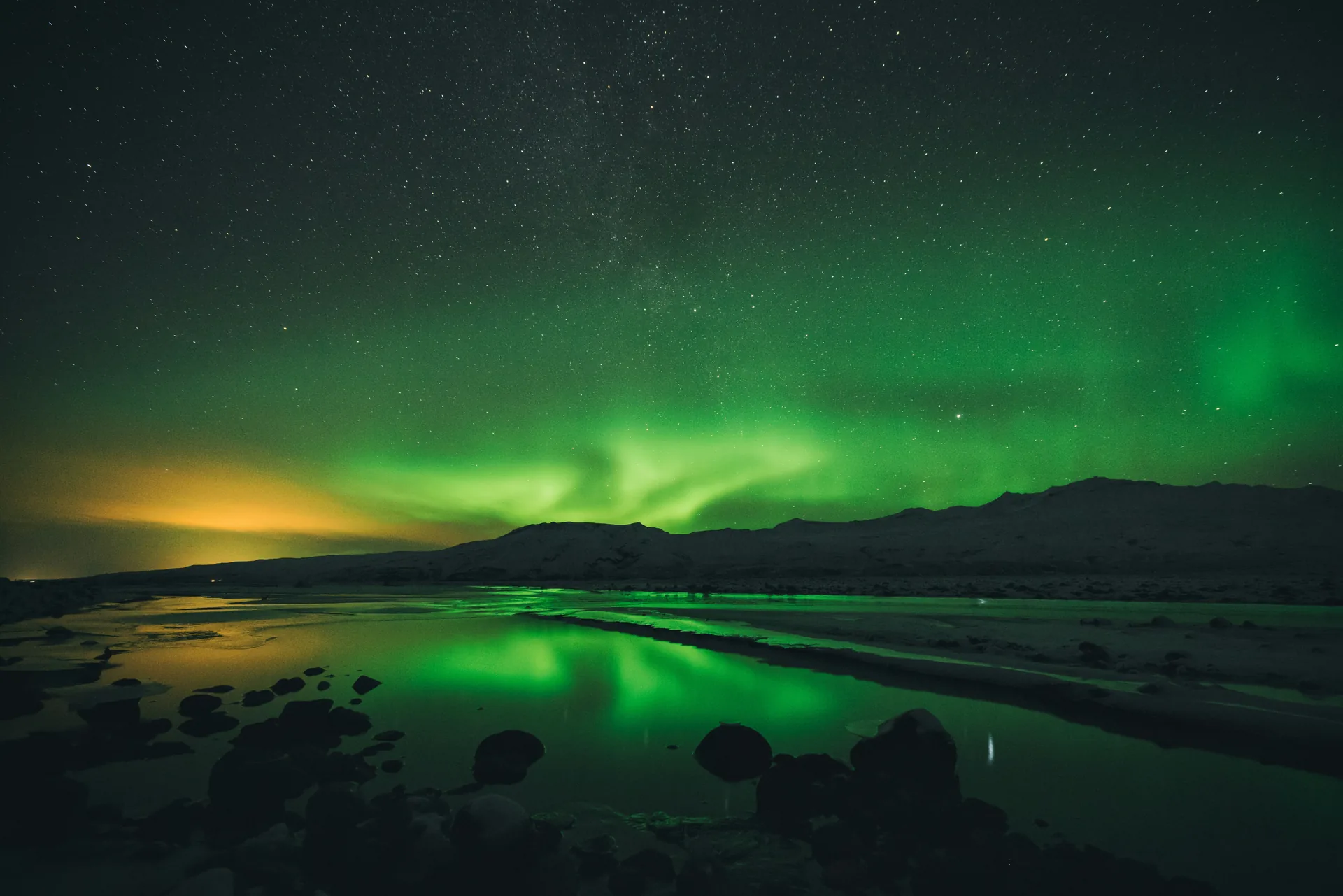 Aurora borealis verde reflectata intr-un lac, cu munti acoperiti de zapada si cer instelat in fundal. Foto: Jonatan Pie / Unsplash