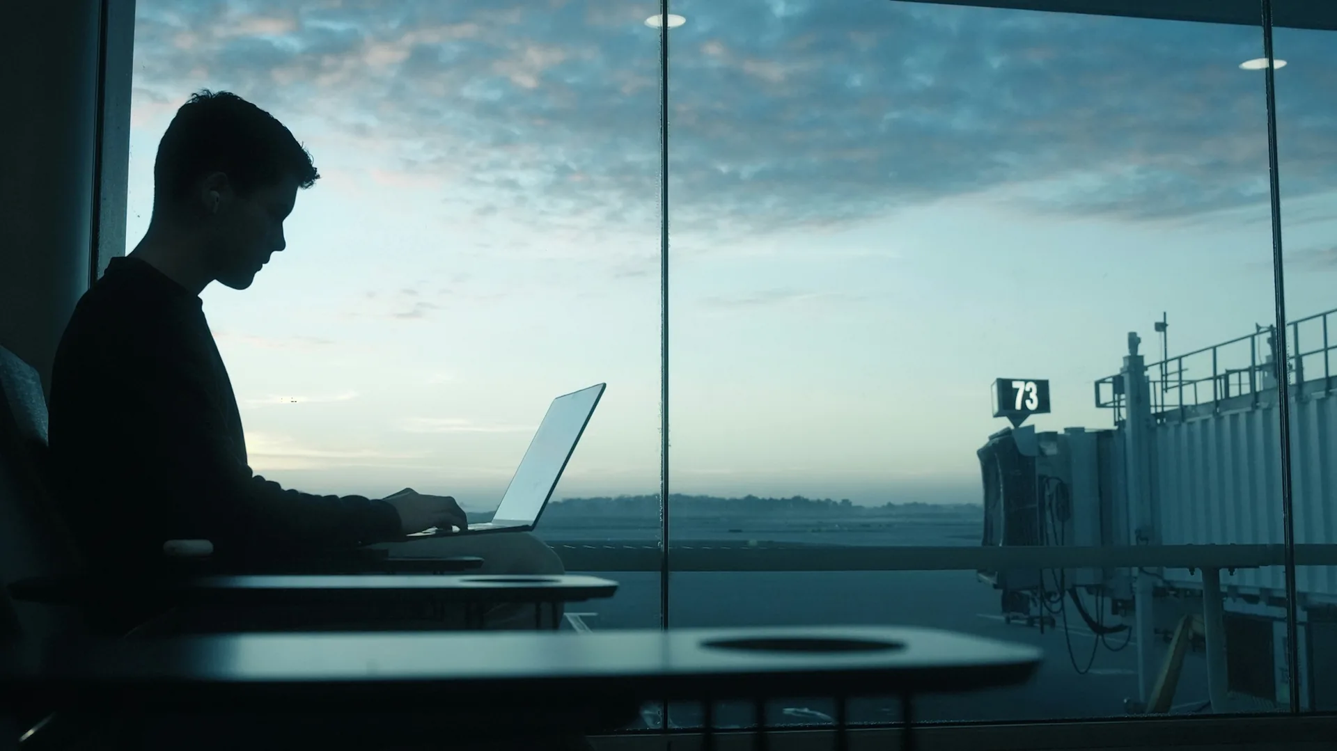 Silueta unui calator lucrand pe laptop la poarta 73 a unui aeroport, la amurg. Foto: Daniel Gomez / Unsplash