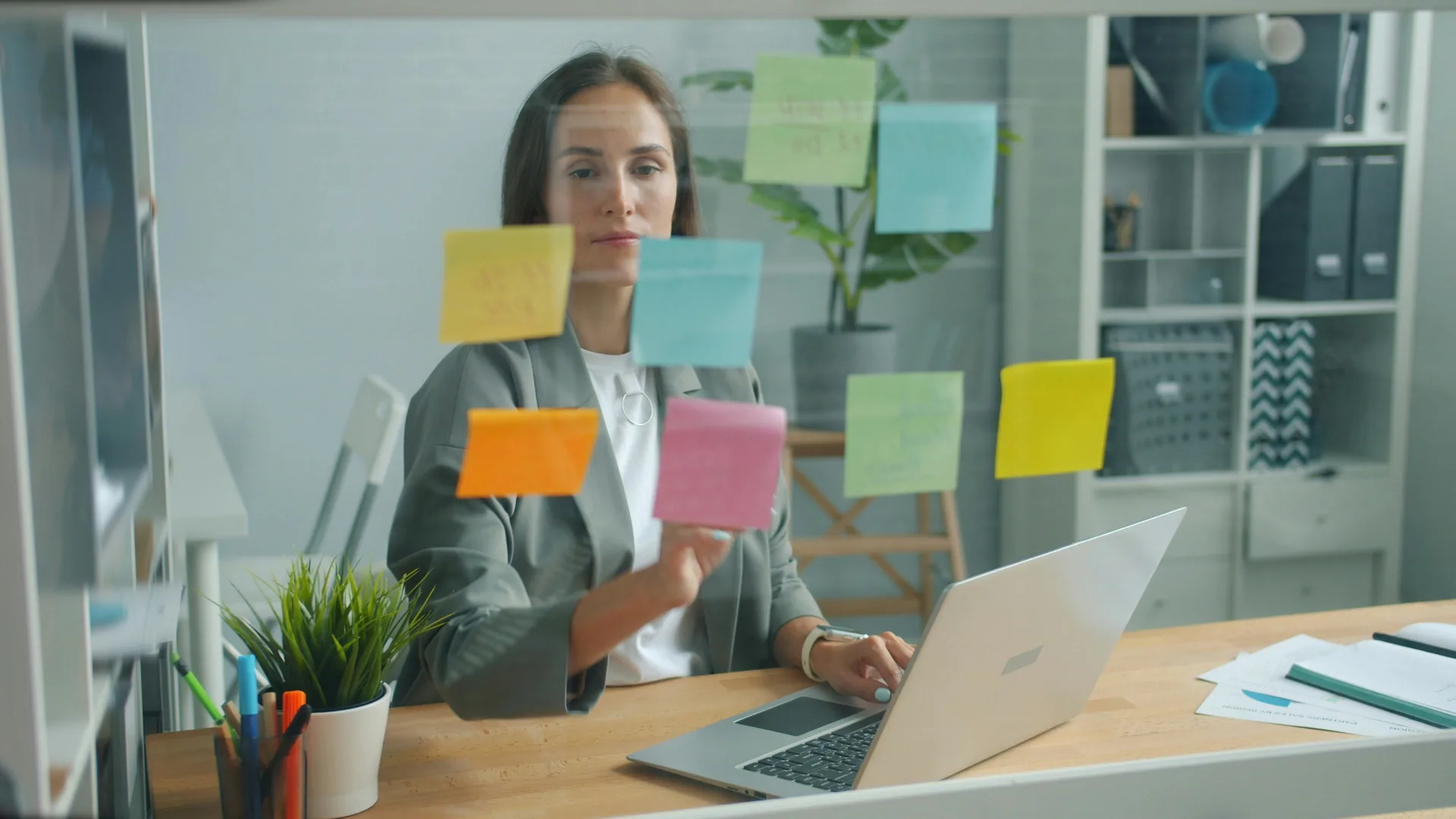 Femeie de afaceri la birou cu laptop, organizand sticky notes colorate pe un panou de sticla. Foto: Vitaly Gariev / Unsplash
