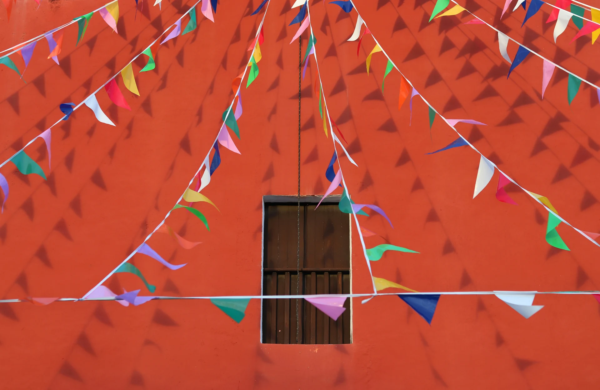 Ghirlande de fanioane colorate in fata unei fatade rosii cu fereastra din lemn, in Mexic. Foto: Analuisa Gamboa / Unsplash