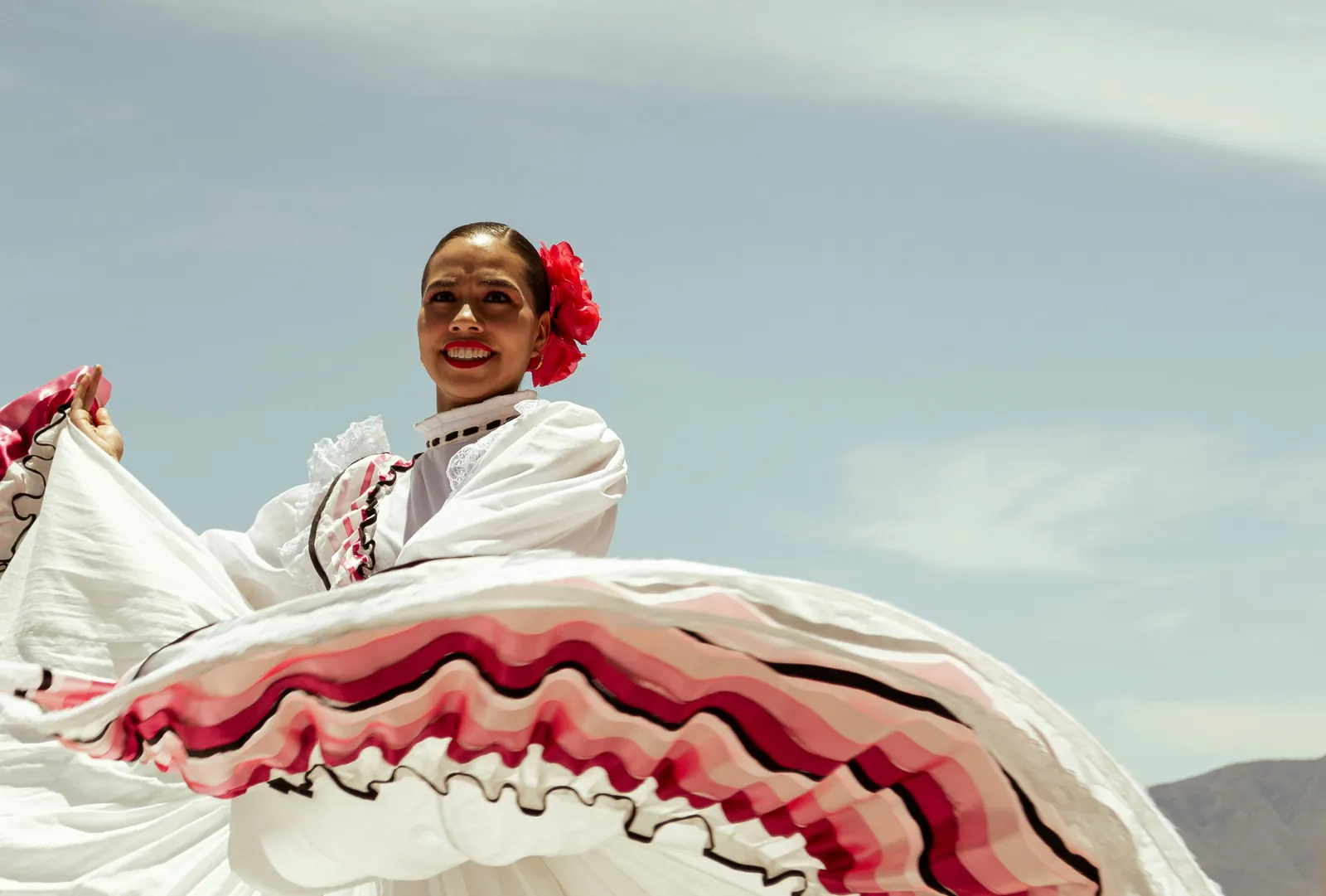 Dansatoare de folclor mexican in rochie alba cu volane roz si negre, rotind fusta pe fundal de cer. Foto: Daniel Pichardo / Unsplash