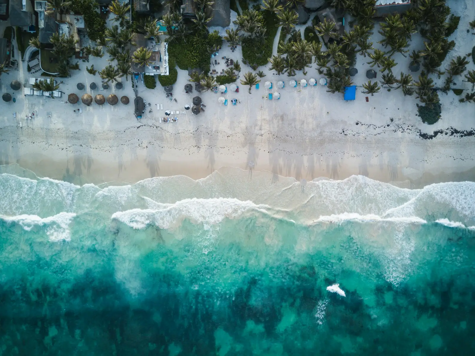 Vedere aeriana asupra unei plaje din Tulum, cu nisip alb, palmieri, umbrele din stuf si apa turcoaz. Foto: Spencer Watson / Unsplash