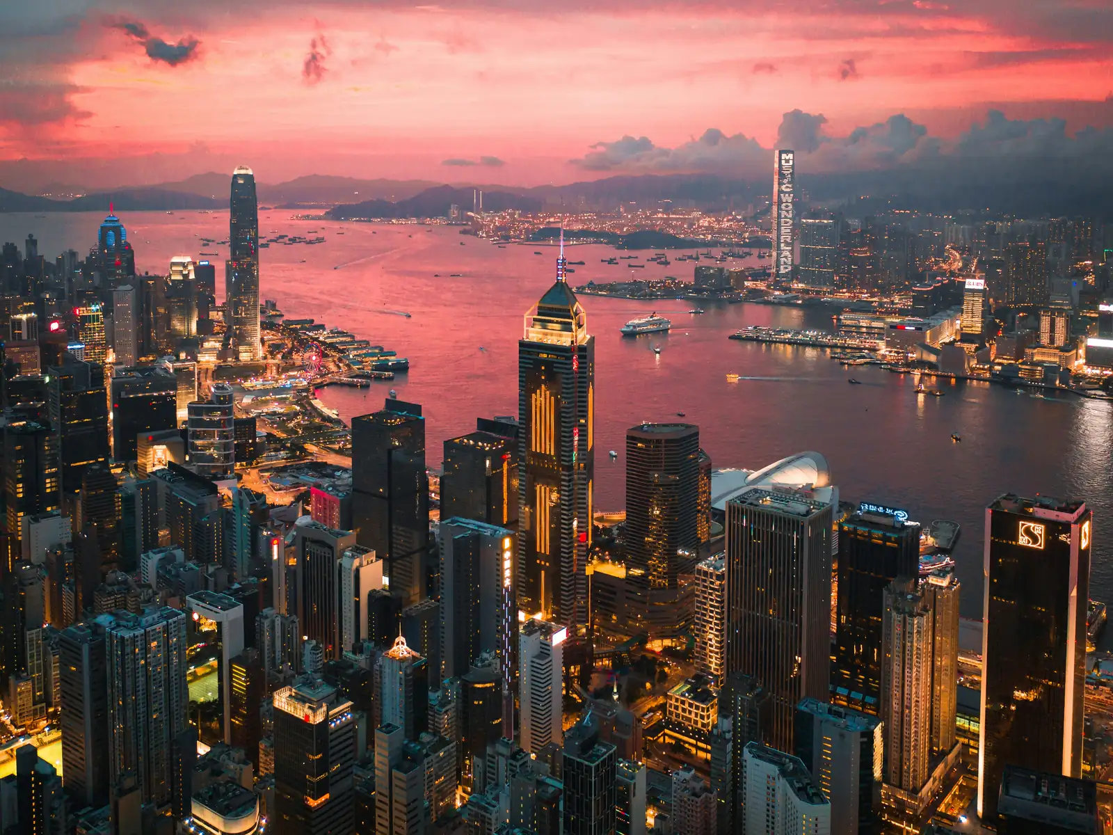 Zgarie-nori din Hong Kong si Portul Victoria vazute de sus, cu luminile orasului aprinse sub un cer roz. Foto: manson / Unsplash