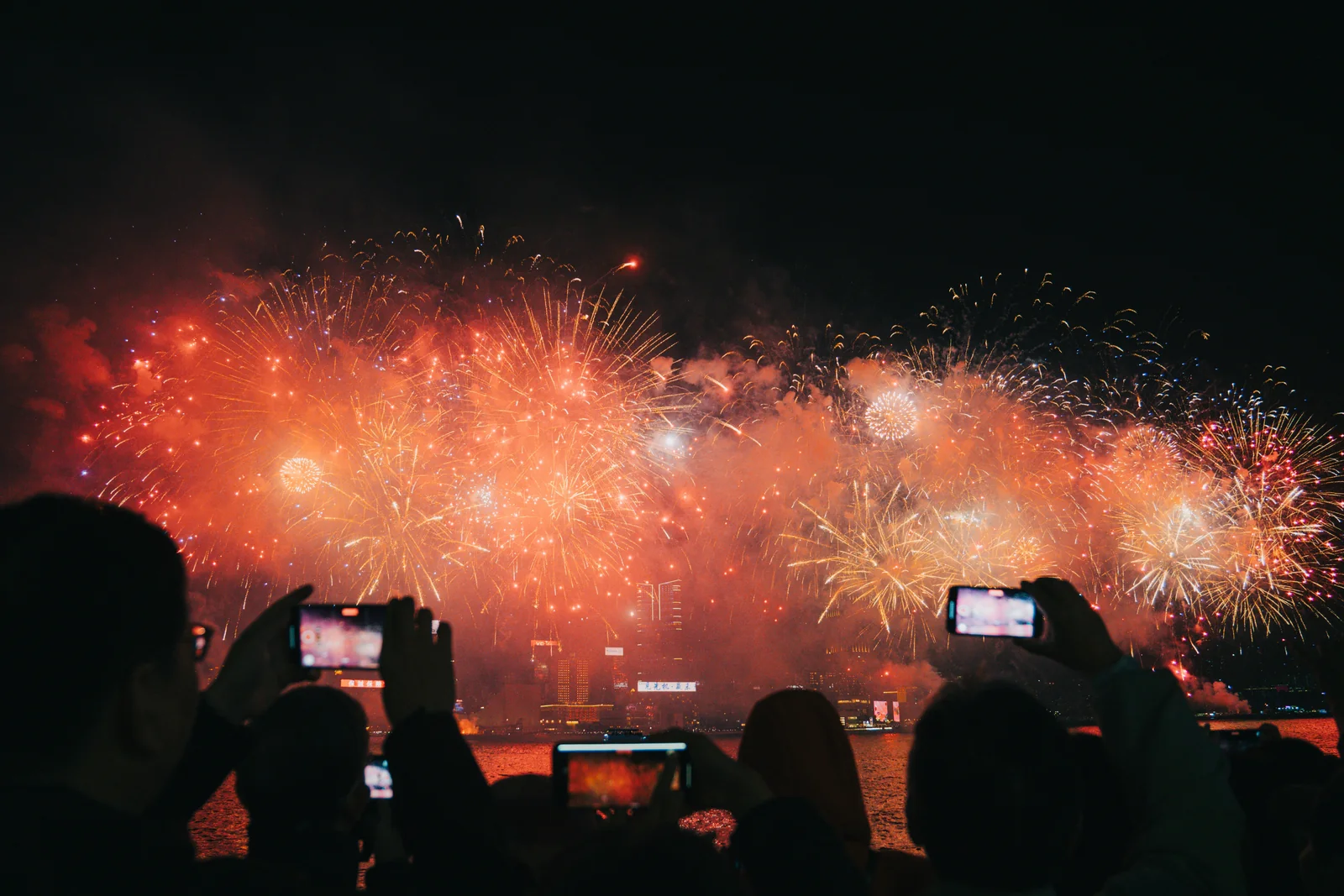 Oameni care filmeaza cu telefonul artificiile de Anul Nou Chinezesc deasupra unui port si a unui oras luminat. Foto: Darren Budiman / Unsplash