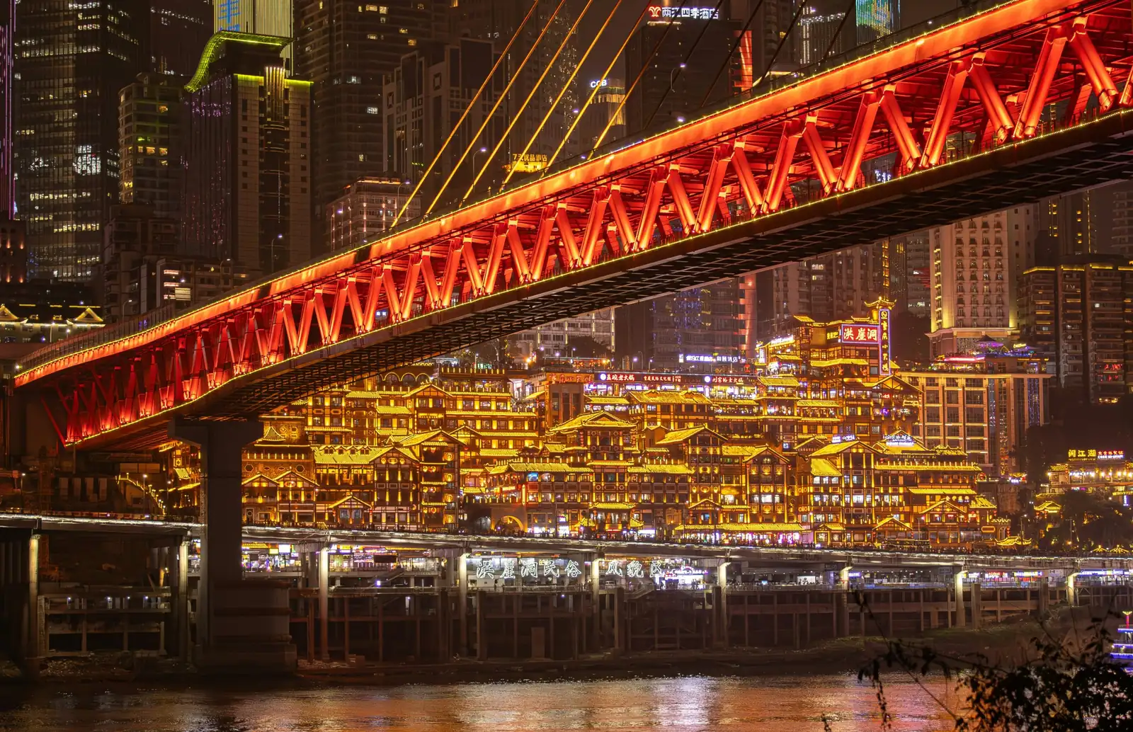 Complexul Hongyadong din Chongqing luminat noaptea, vazut de peste rau, sub podul Qiansimen iluminat in rosu. Foto: Irvin Liang / Unsplash