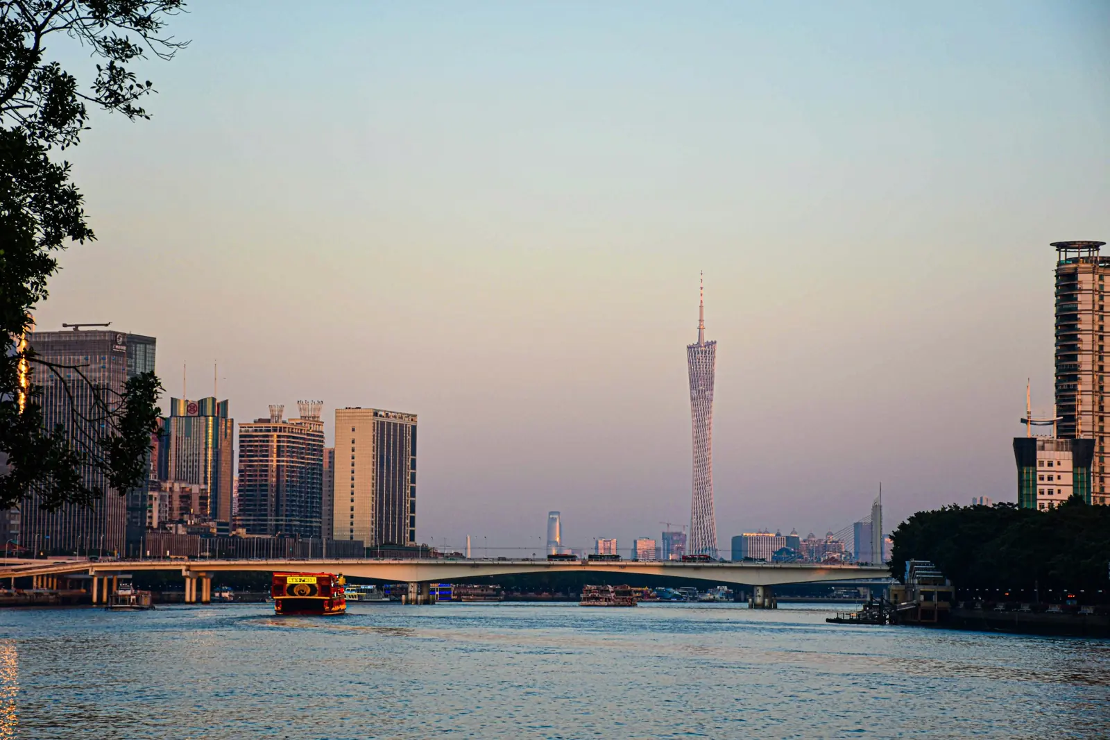 Turnul Canton si zgarie-nori din Guangzhou deasupra Raului Perlelor, cu un vapor turistic rosu, in lumina calda. Foto: Lan Lin / Unsplash