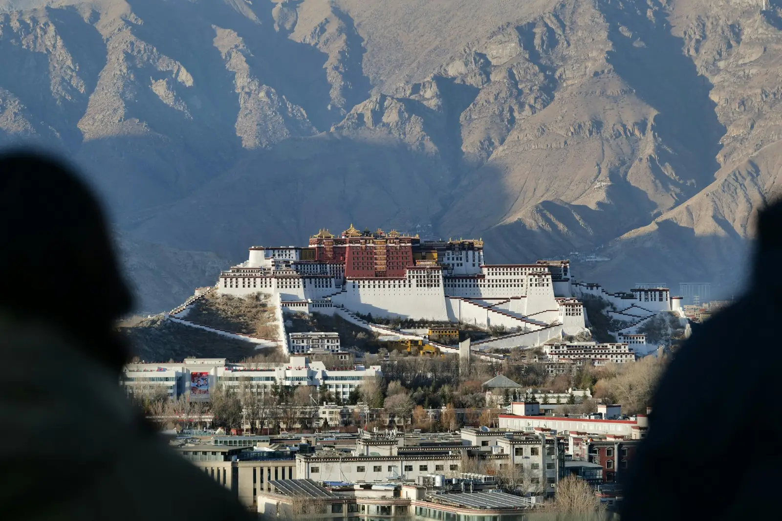 Palatul Potala din Lhasa, cu zidurile albe si rosii luminate de soare, vazut printre siluete, cu muntii in fundal. Foto: Xuyu Chi / Unsplash
