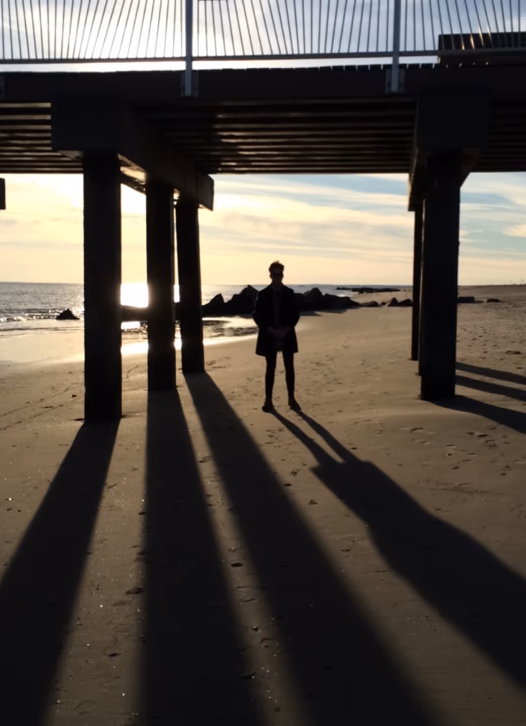 Person standing on a beach under a pier at sunset, casting long shadows on the sand.