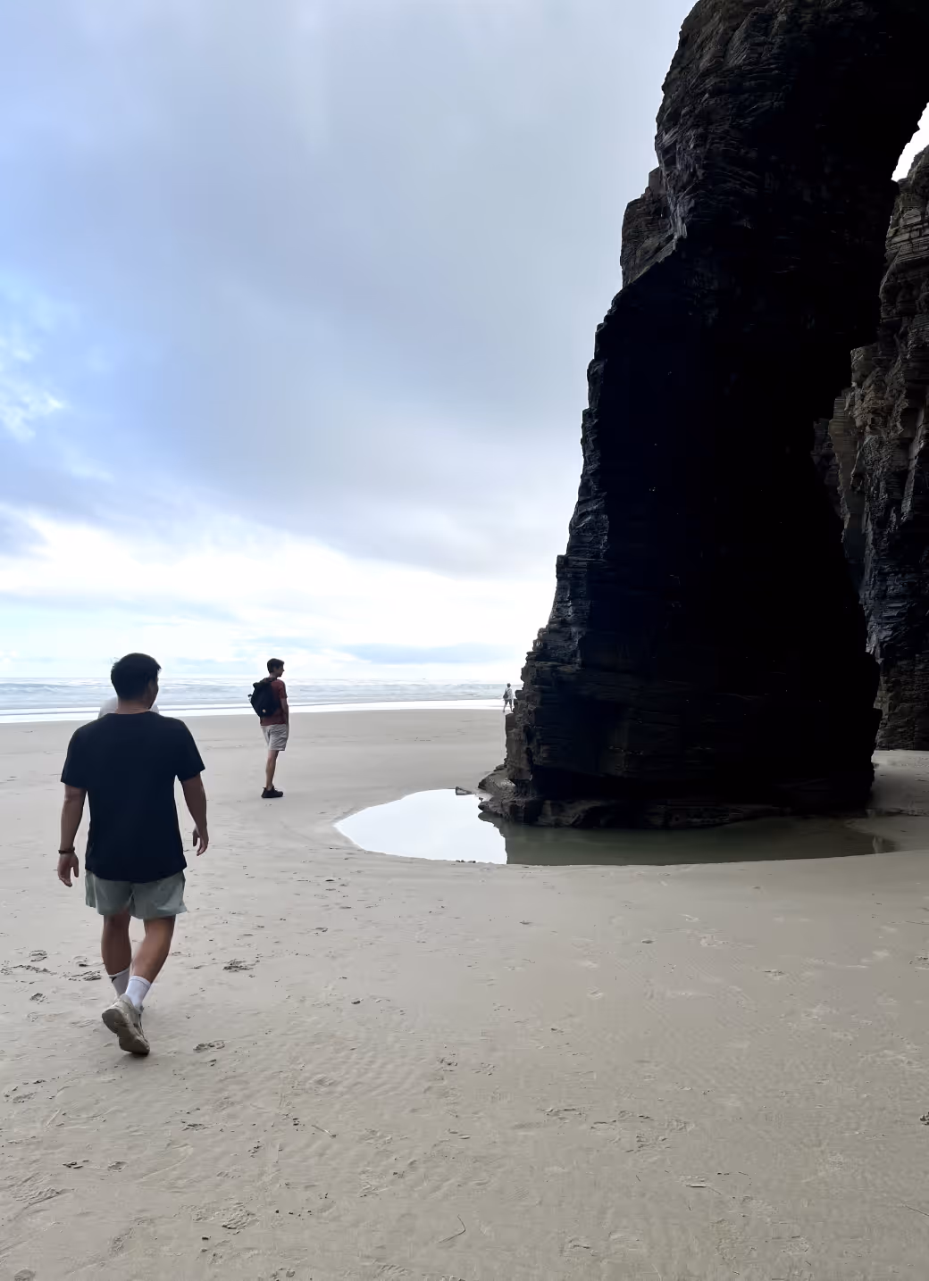 Three people walking on a sandy beach near a large dark rock formation with a shallow pool of water beside it under a cloudy sky.