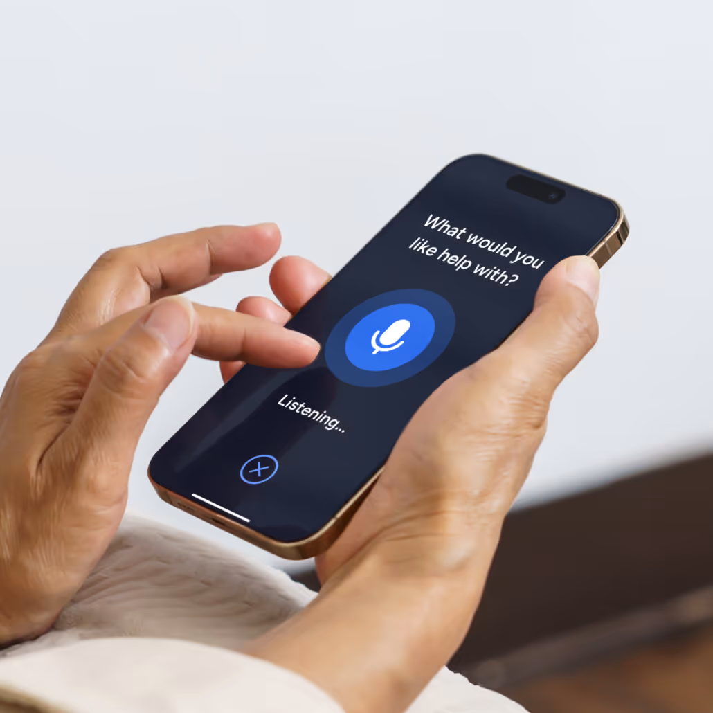 Person holding a smartphone with a voice assistant screen displaying the prompt 'What would you like help with?' and a blue microphone icon showing 'Listening...'.