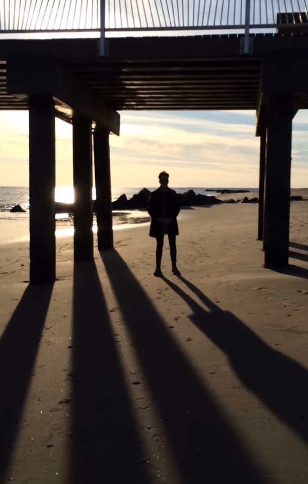 Silhouette of a person standing under a pier on a beach with long shadows cast by the setting sun.