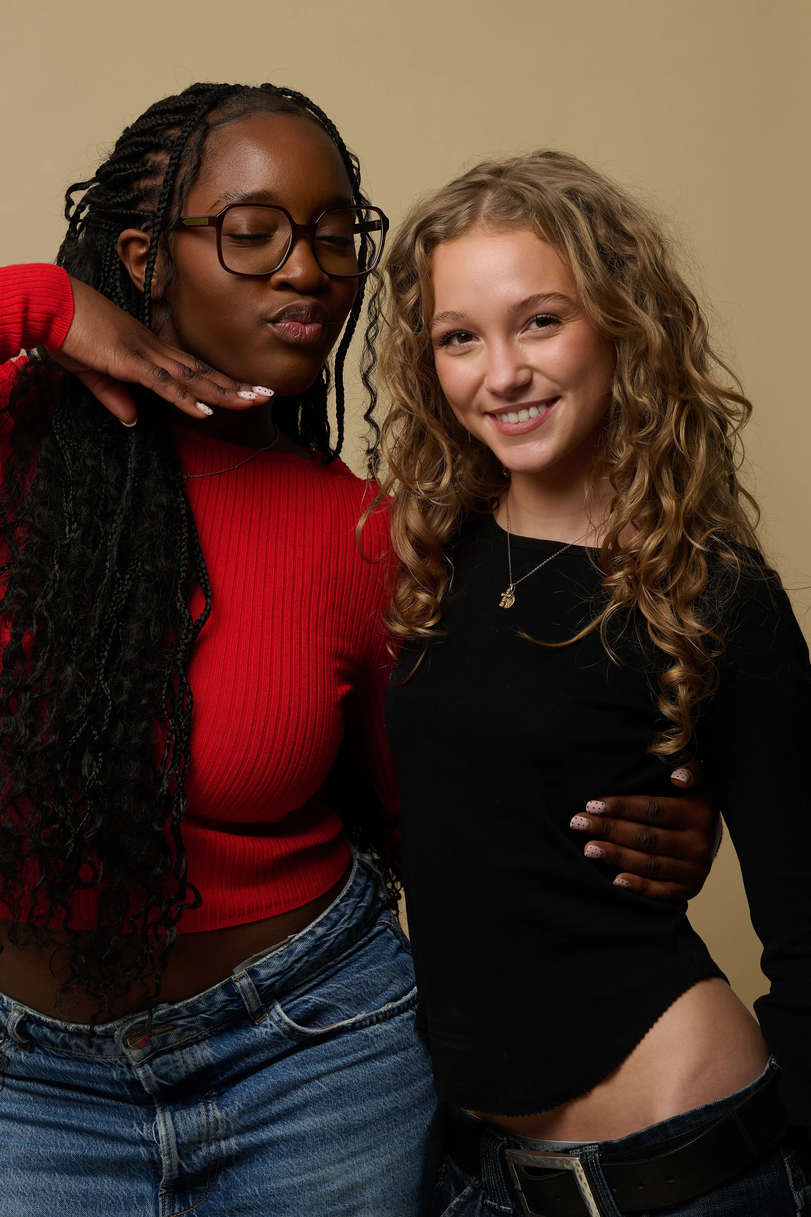 Two young women posing against a beige background, one with long braided hair wearing a red sweater and glasses, the other with curly blonde hair wearing a black top and smiling.