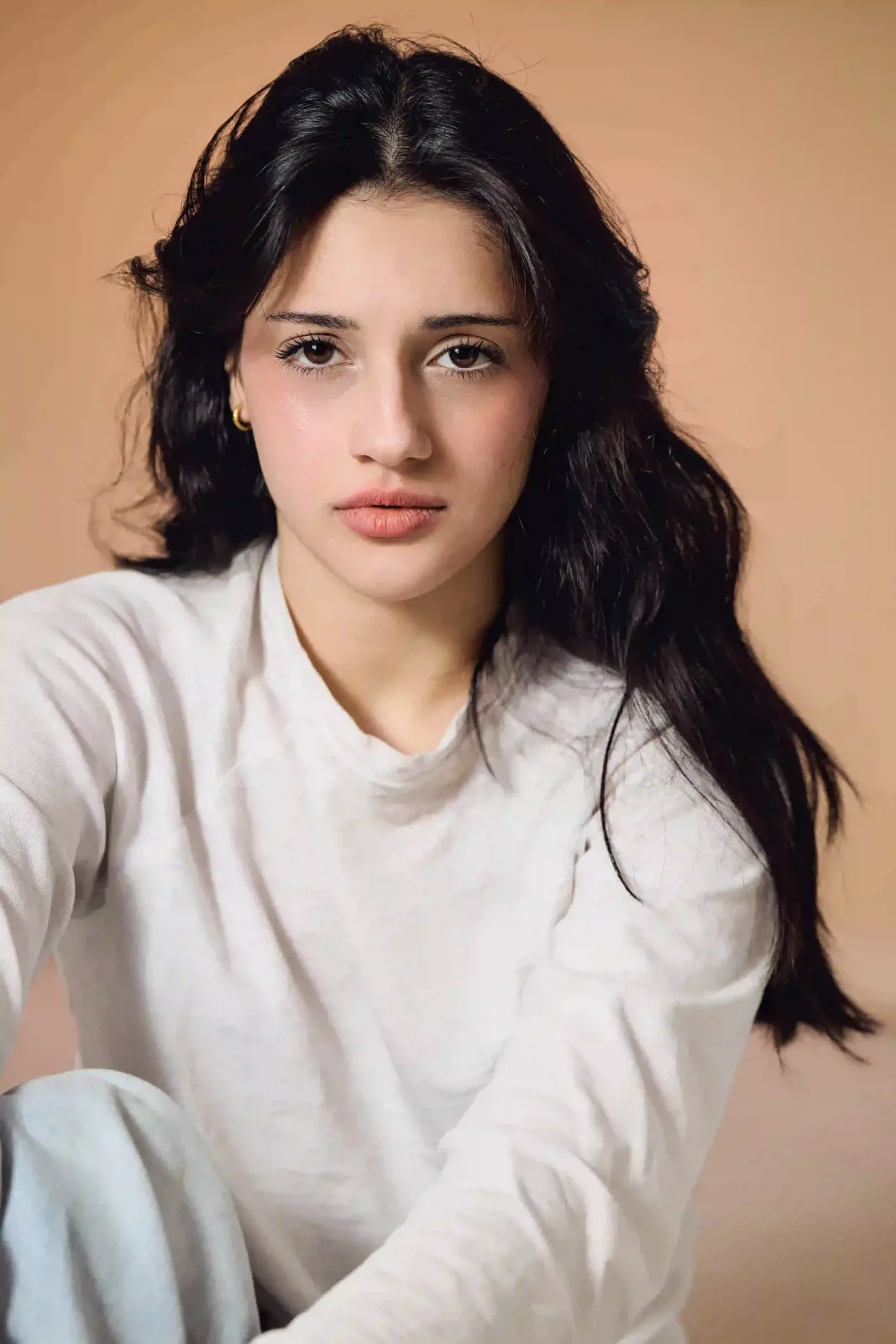 Editorial portrait of actor Nicole Sheyni with long dark hair in a white top and light blue jeans, seated against a warm beige backdrop — actor headshots in Leeds at Dock Street Studio