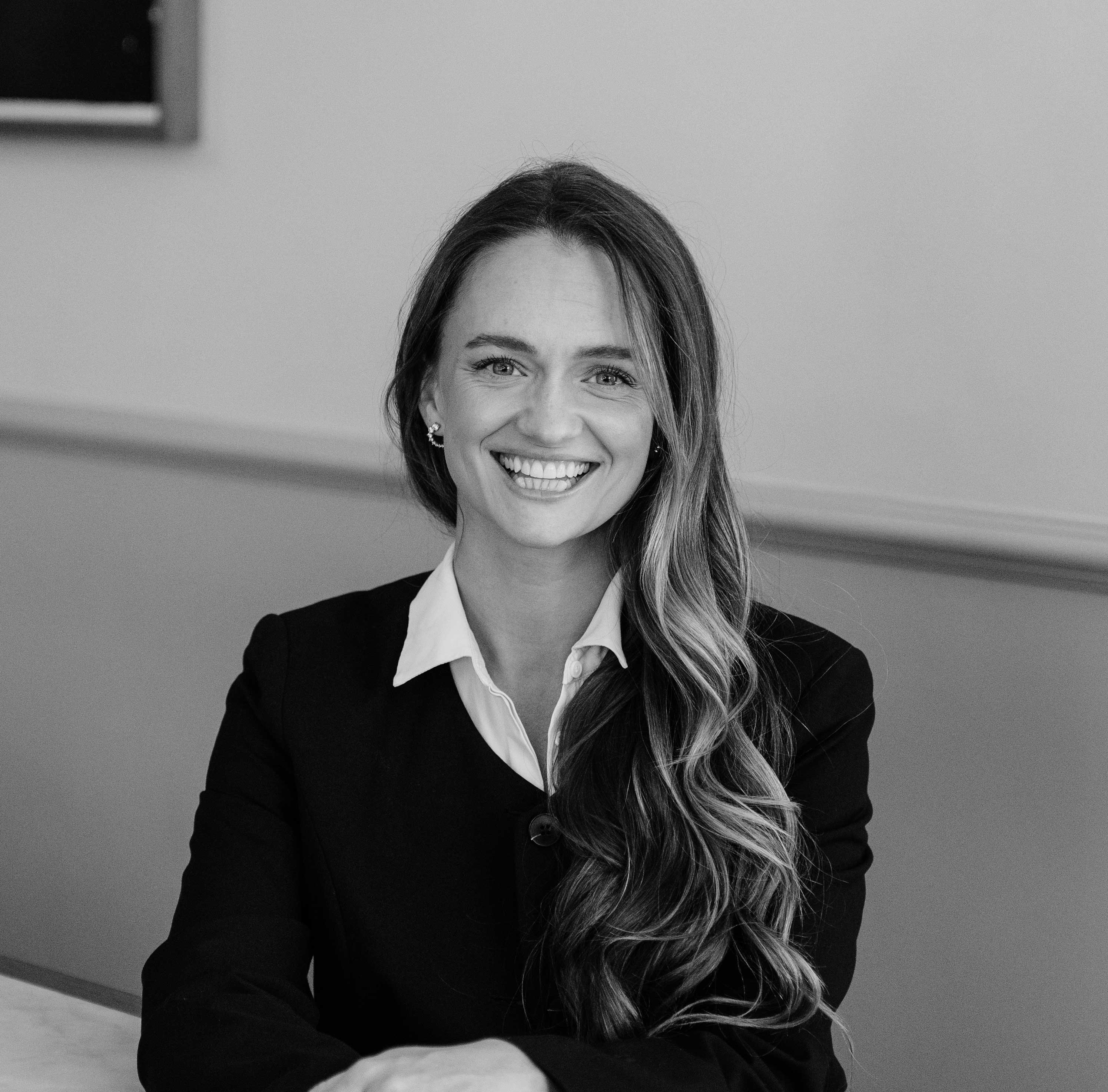 Smiling woman with long wavy hair wearing a dark blazer and white collared shirt in black and white photo.