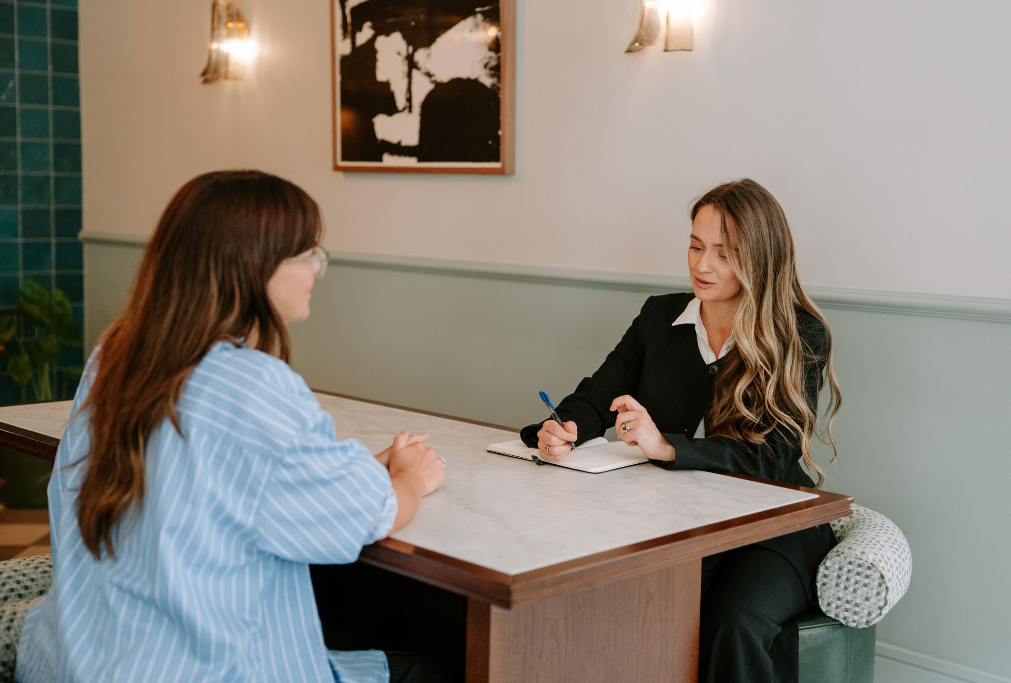 Two women sitting at a table, one writing in a notebook while talking to the other.