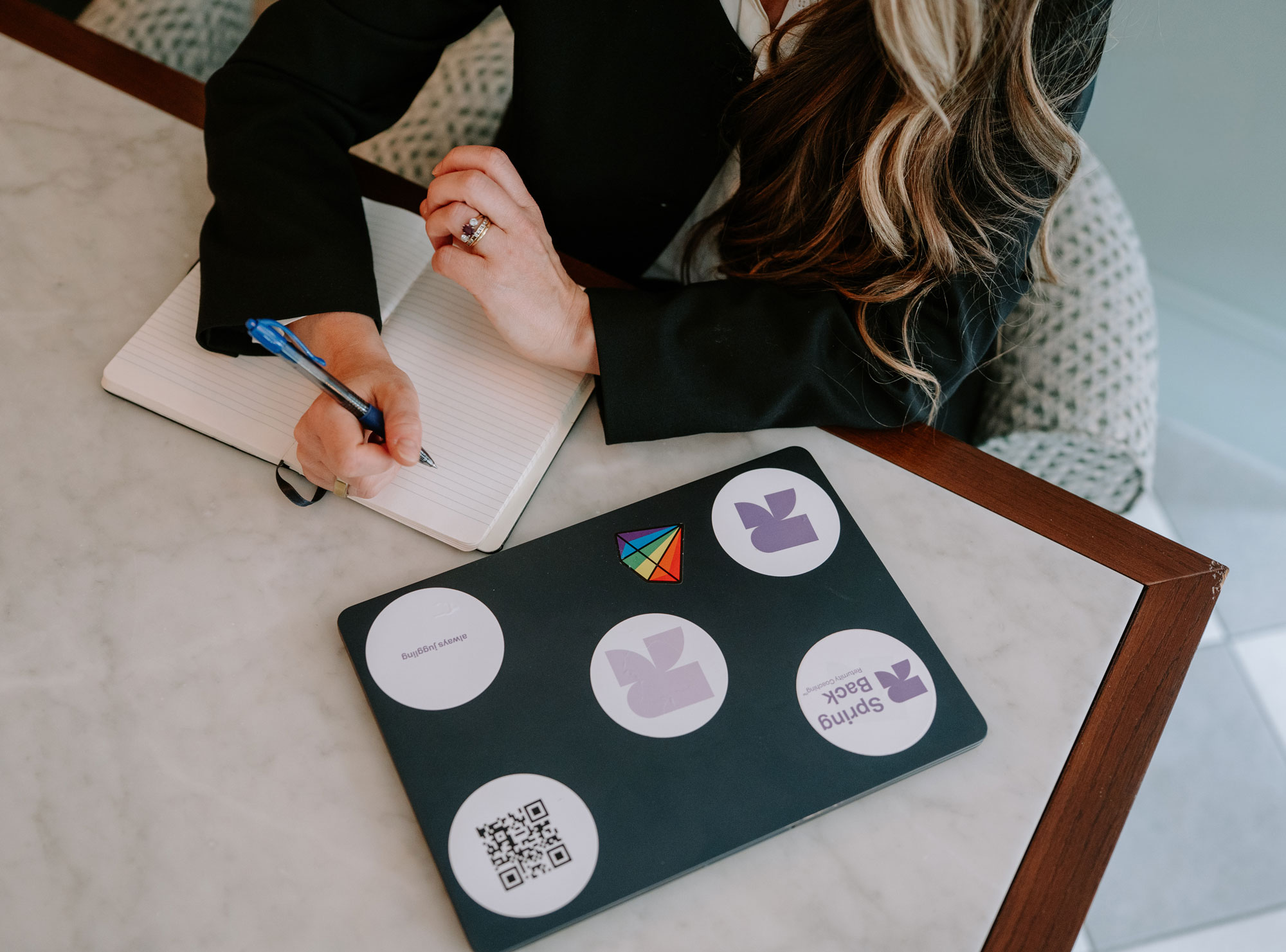 Person in black jacket writing in notebook by laptop with colorful stickers on marble table.