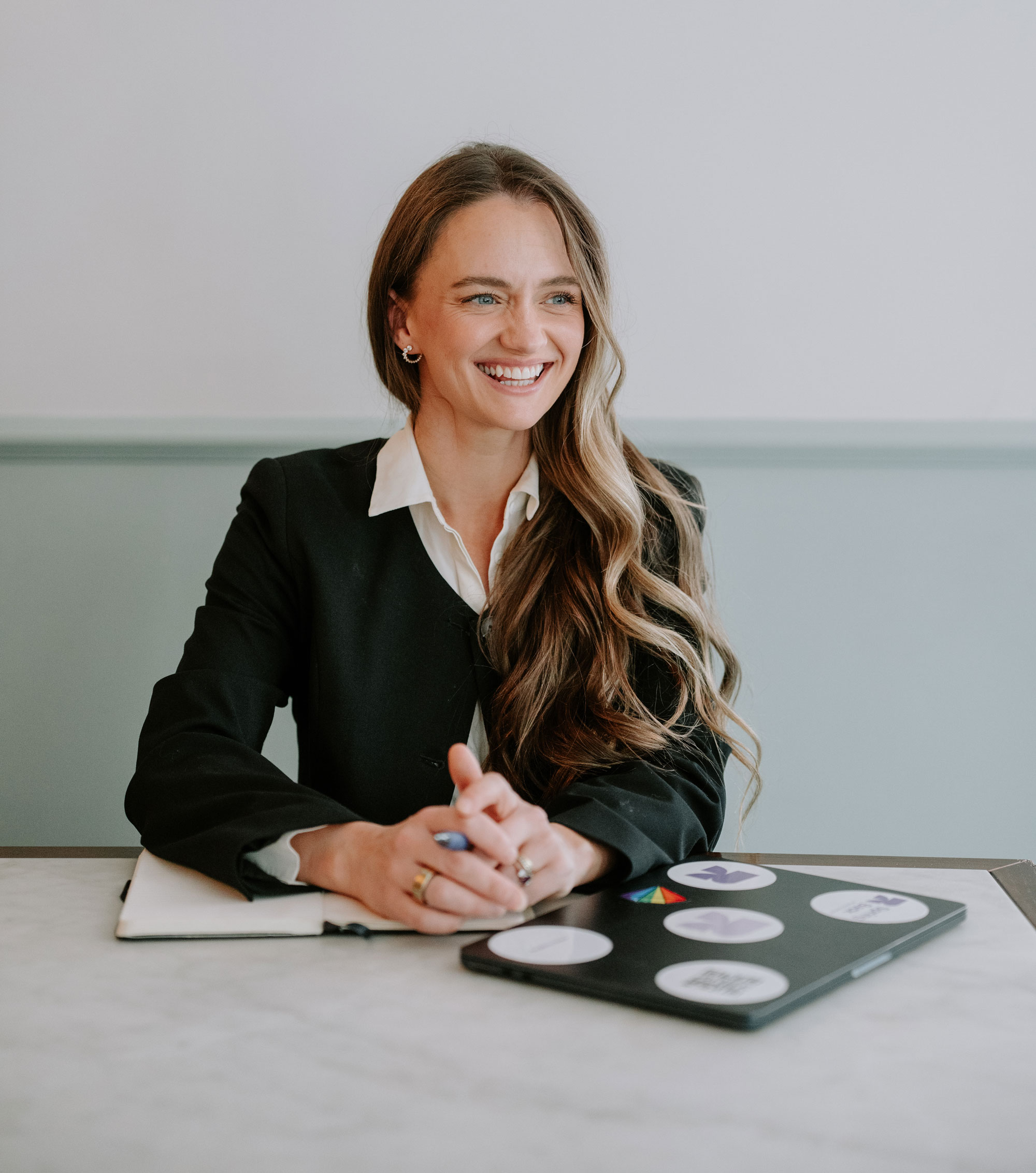Smiling woman with long wavy hair sits at table with laptop covered in round stickers and a notebook.