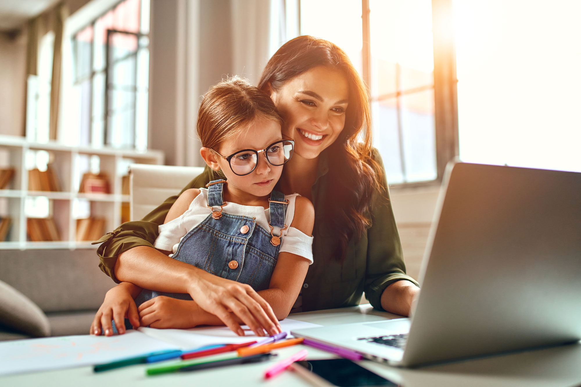 Smiling woman helps young girl with glasses use a laptop at a desk with scattered colored markers.