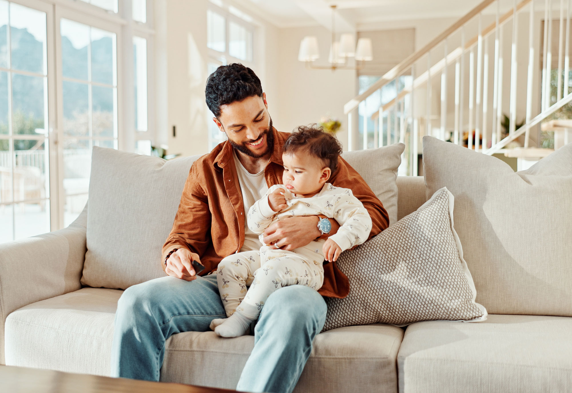 Smiling man holding a baby on his lap while sitting on a beige couch in a bright living room.