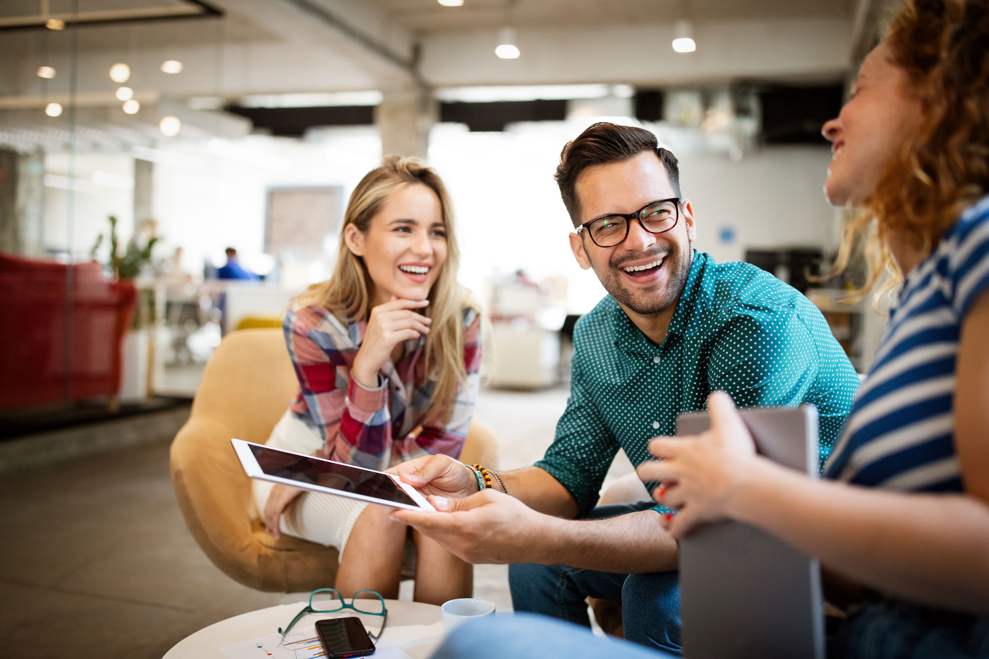 Three colleagues smiling and discussing while sharing a tablet in a modern office lounge.