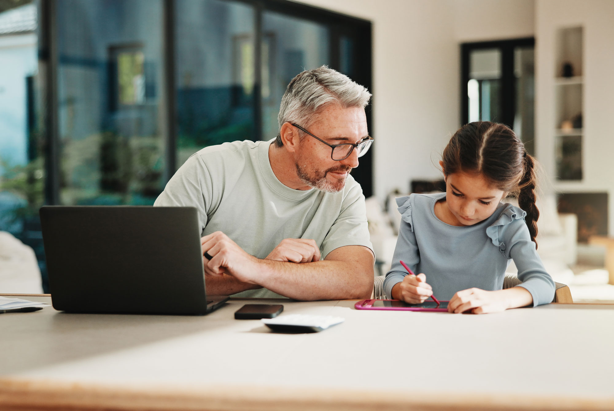Man with glasses watches girl drawing on tablet at table with laptop and phone nearby.