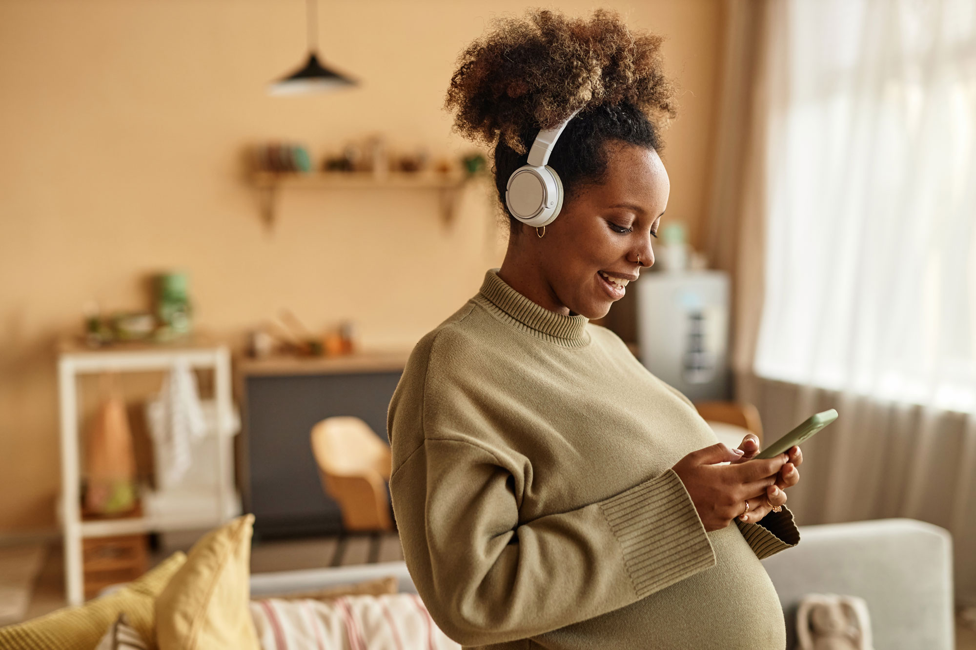 Pregnant woman in headphones smiling and using smartphone in a cozy living room.