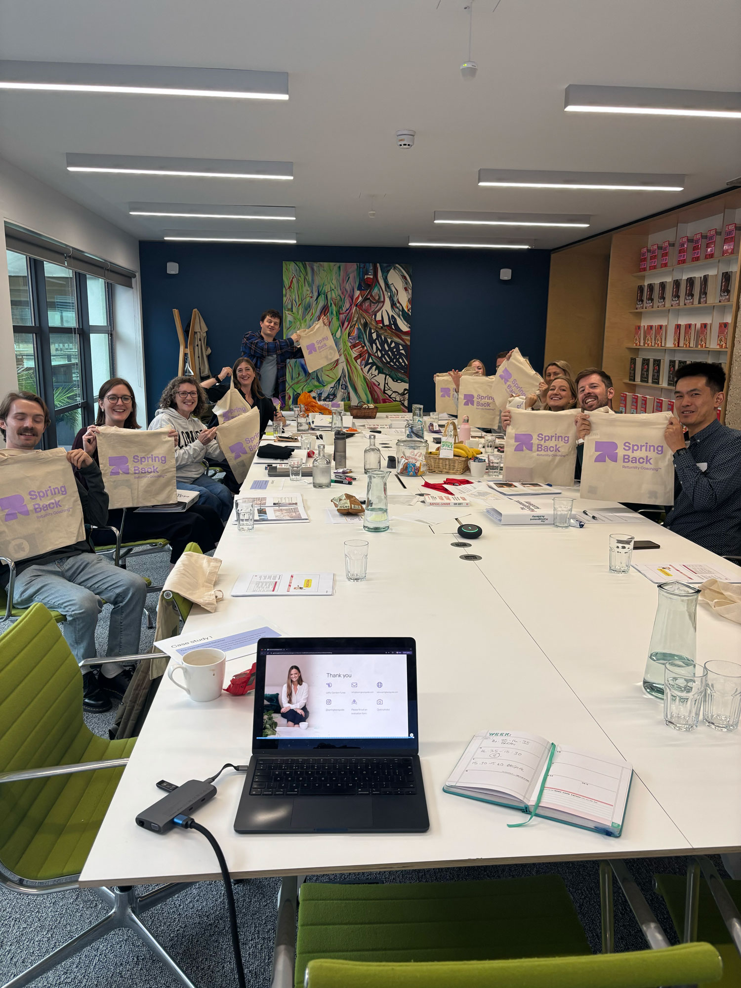 Group of people around a large table holding up beige tote bags with purple Spring Back logos.