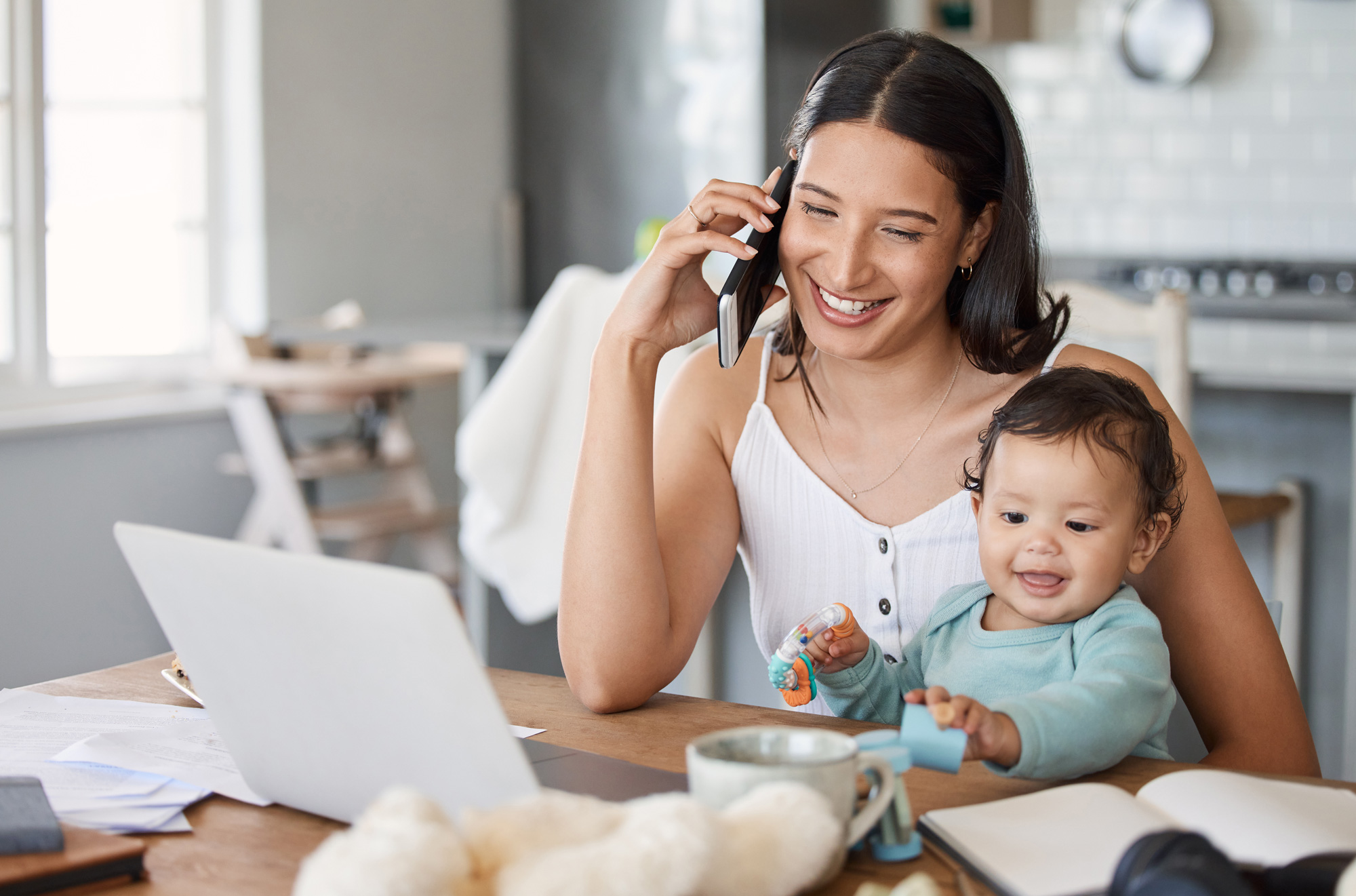 Smiling woman talking on phone while holding a baby playing with toys at a table with a laptop.