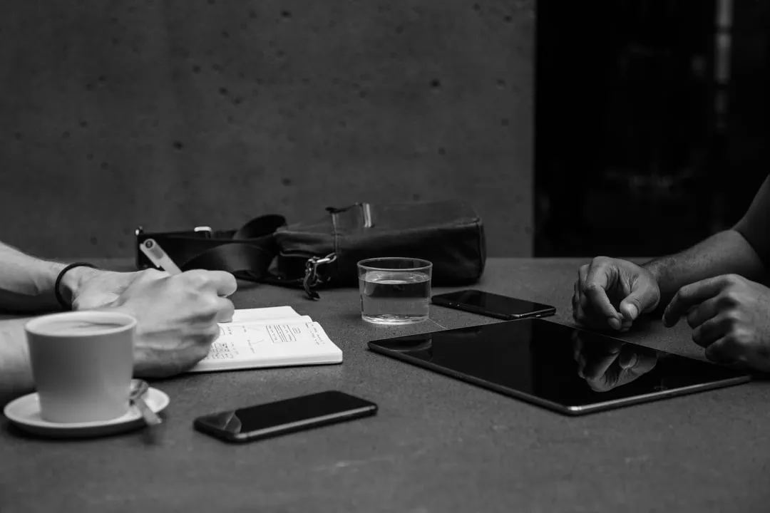 A man is writing in a notebook while a tablet is on the table.