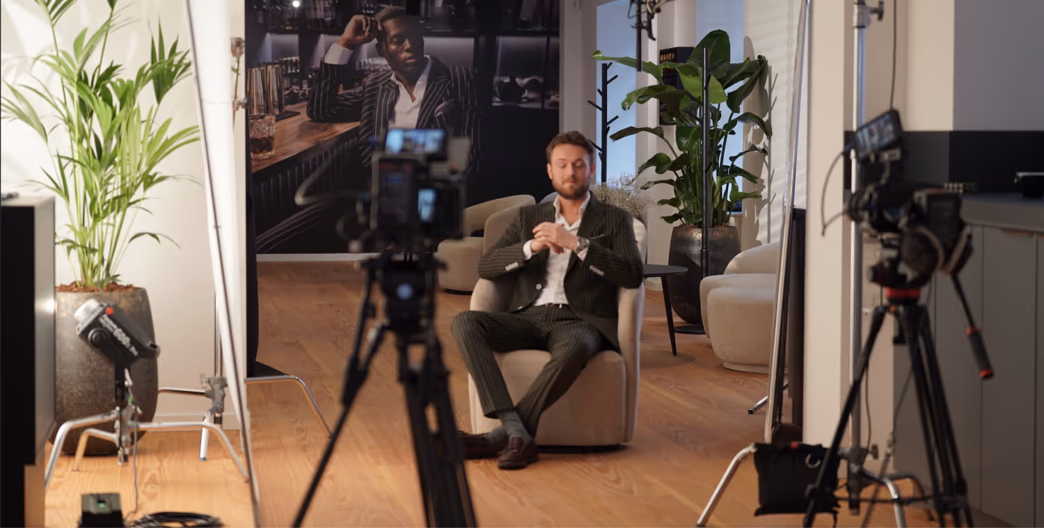 Mand i jakkesæt sidder i en stol under optagelse med kameraer og planter i baggrunden.