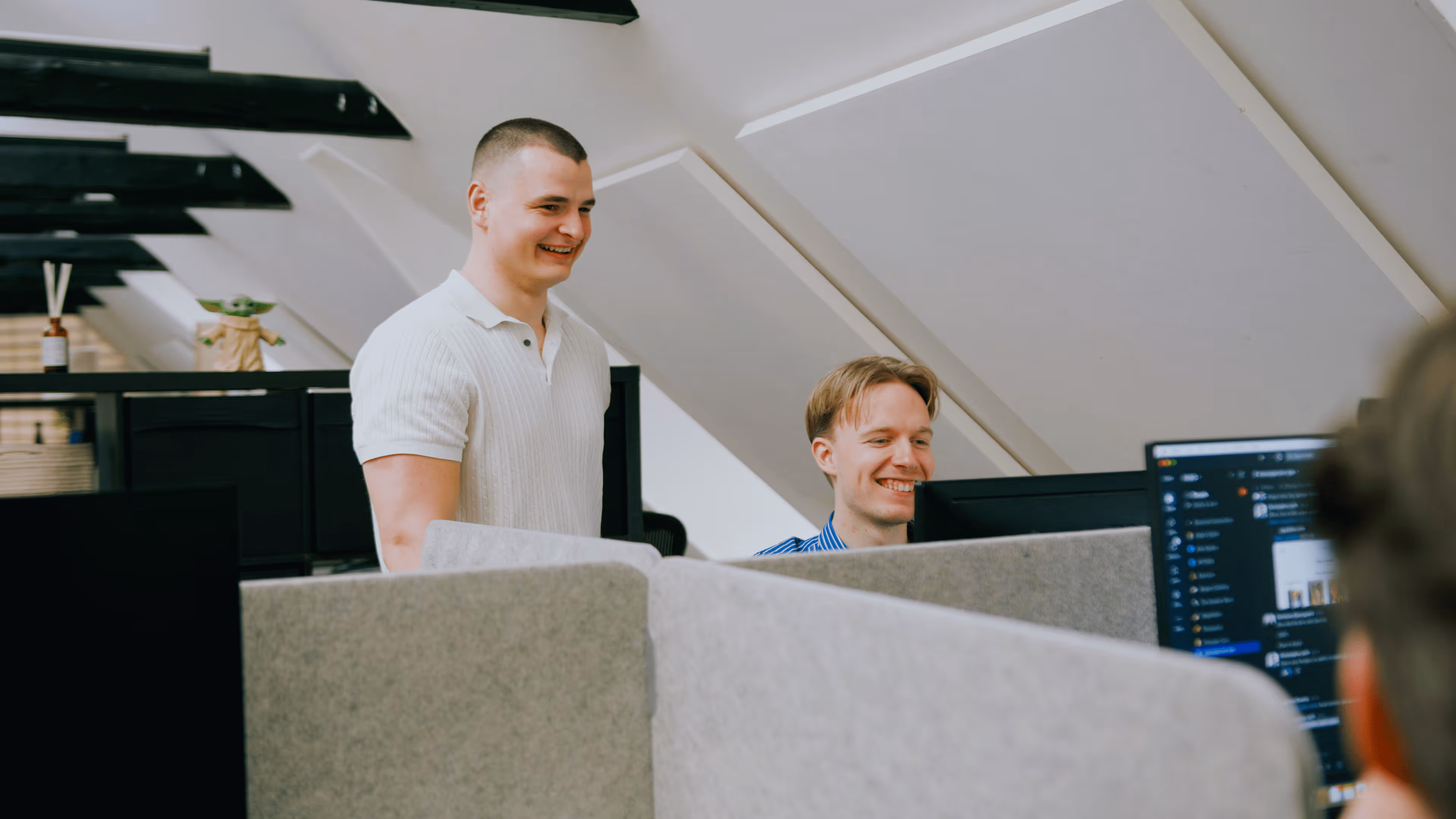 To mænd smilende og samarbejdende ved kontorcomputere i et åbent kontormiljø med skrå lofter.