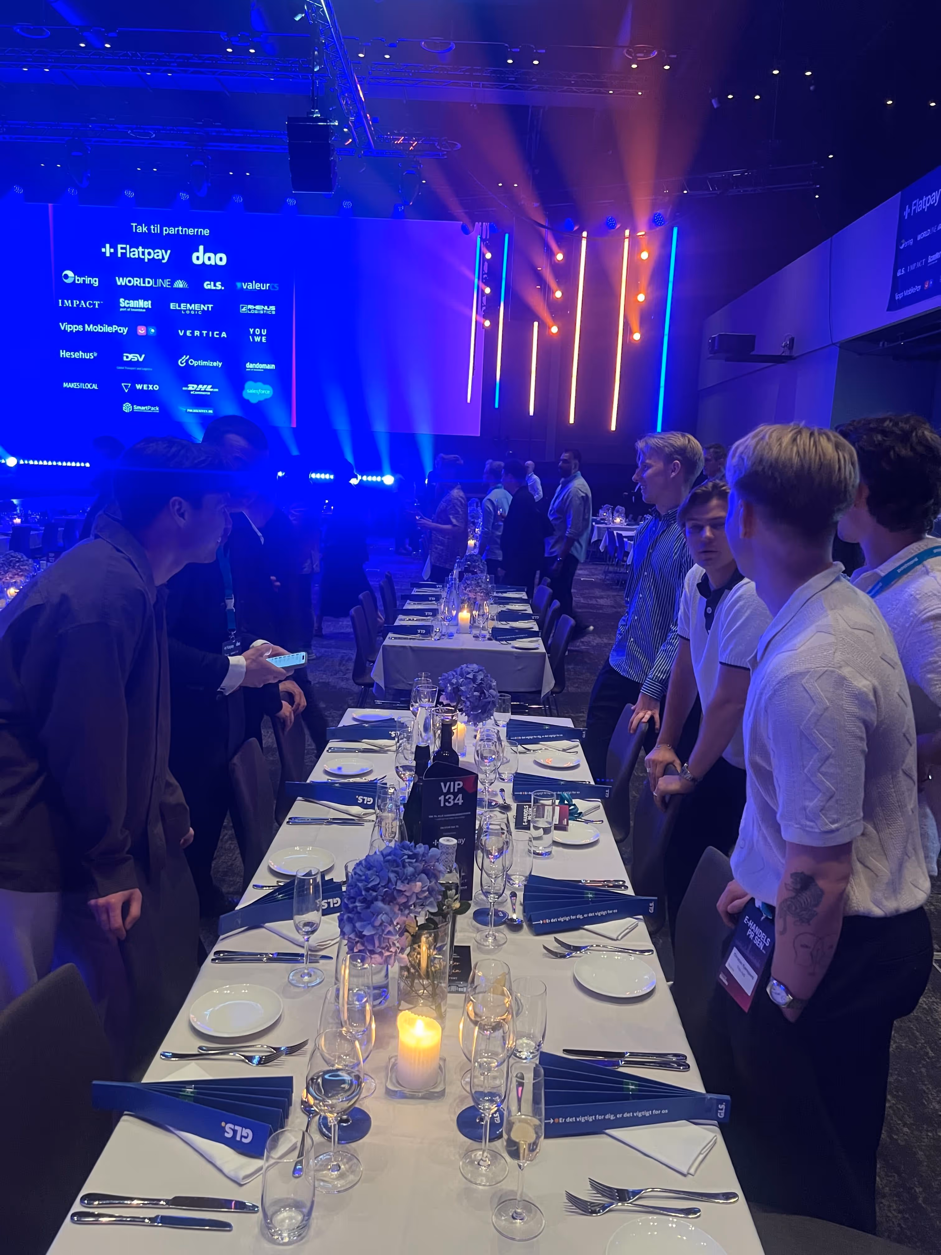 Gæster står op ved et langbord dækket med glas, tallerkener og blomster under et oplyst event med blå og orange lys.
