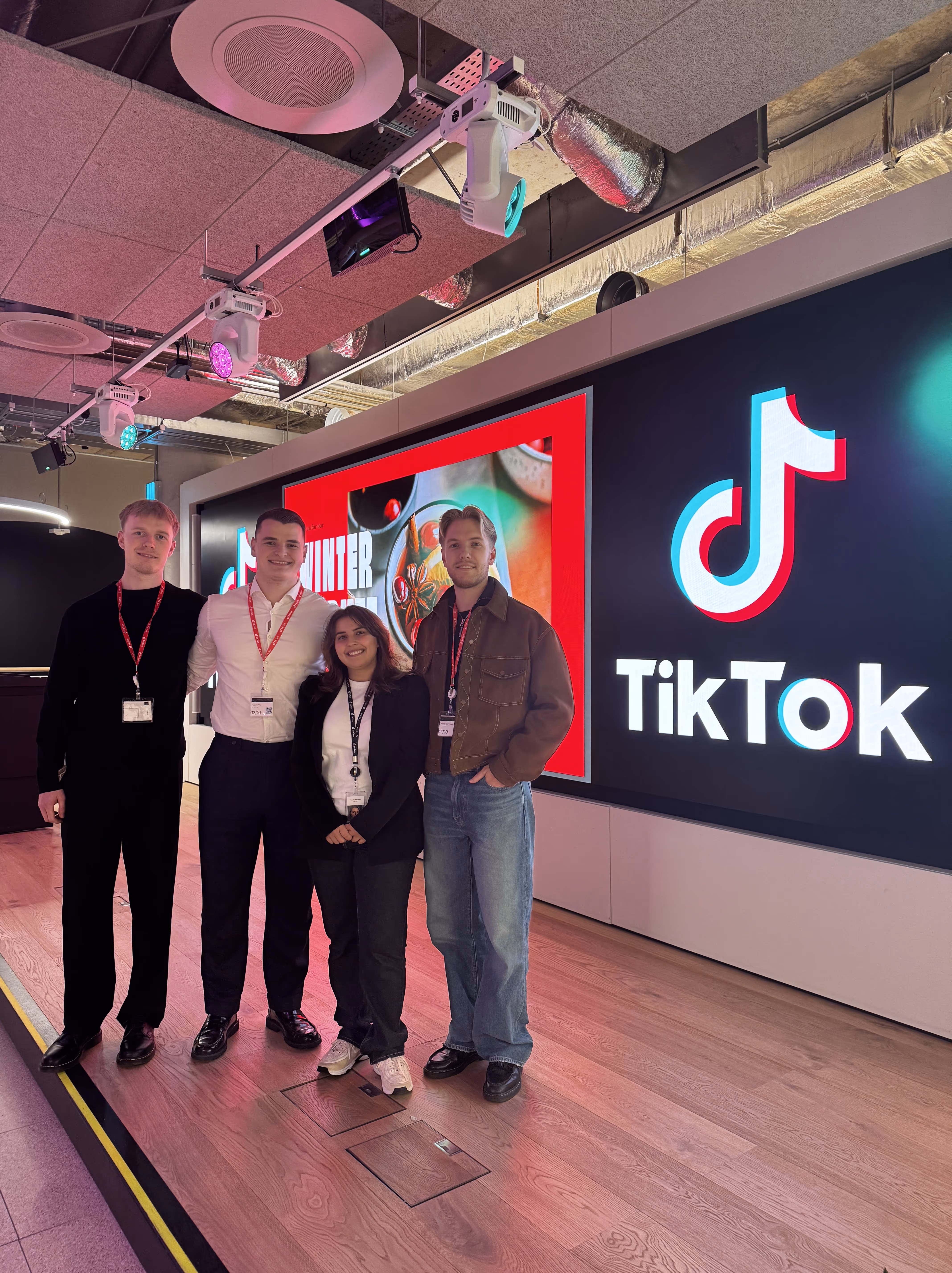 Fire unge voksne står foran en stor TikTok-skærm i et moderne kontormiljø.
