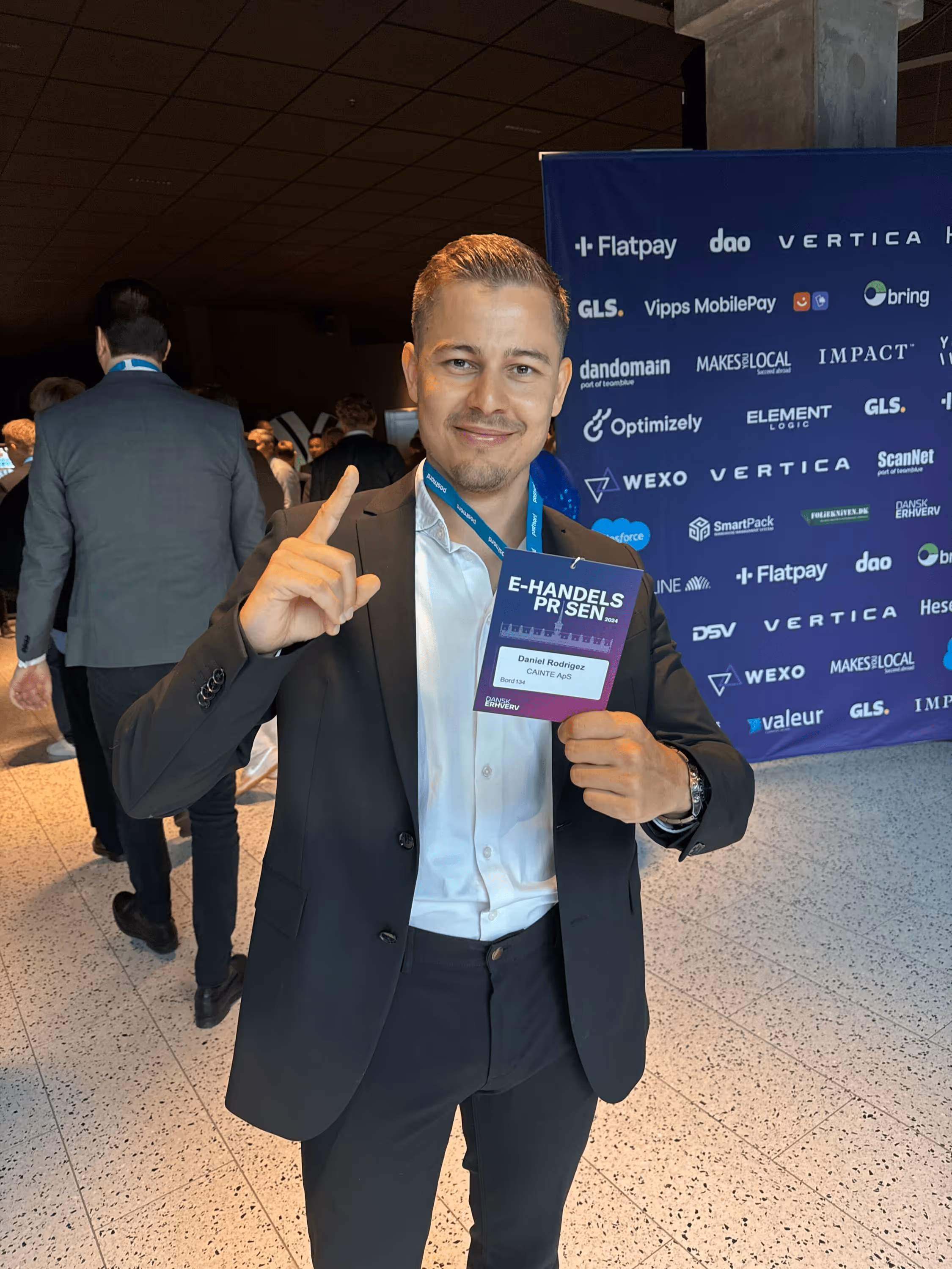 Mand i sort jakkesæt smilende og holder badge med teksten 'E-HANDELSPRISEN 2024' ved en event med sponsor-logoer i baggrunden.