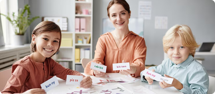 Una maestra y dos niños sentados en una mesa, usando tarjetas para aprender palabras.