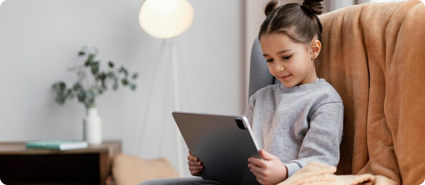 A young girl sitting on a chair, using a tablet to learn new English words in a fun way.