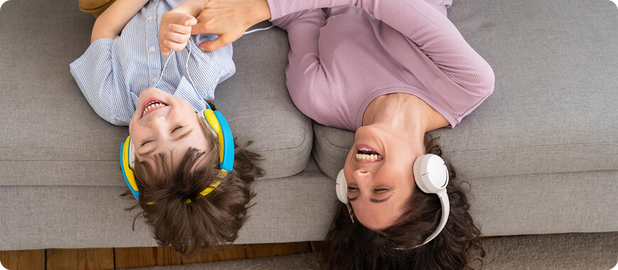 A mother and child lying on a couch, both wearing headphones and smiling while practicing English listening activities together.