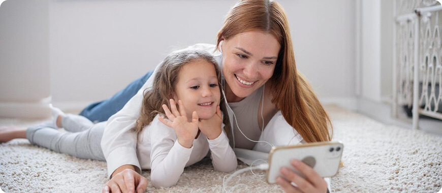 A mother and daughter lying on the floor, using earphones and a smartphone to engage in an English listening activity.