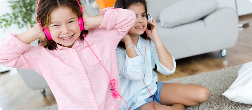 Two young girls sitting on the floor, wearing colorful headphones and smiling while enjoying an English listening activity.