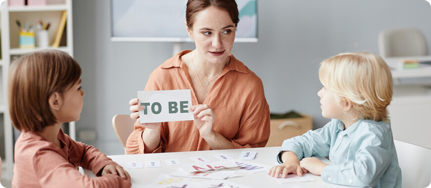 Maestra o mamá enseñando a niños el verbo 'to be' con tarjetas – actividad interactiva y divertida de gramática inglesa