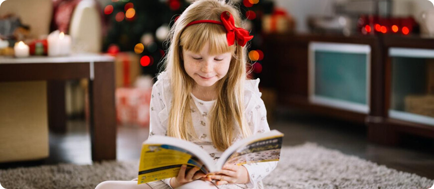 Learn Adjectives Through Storytime Little girl reading book near Christmas tree — perfect setting to learn descriptive adjectives like ‘cozy’, ‘sparkly’, ‘festive’.