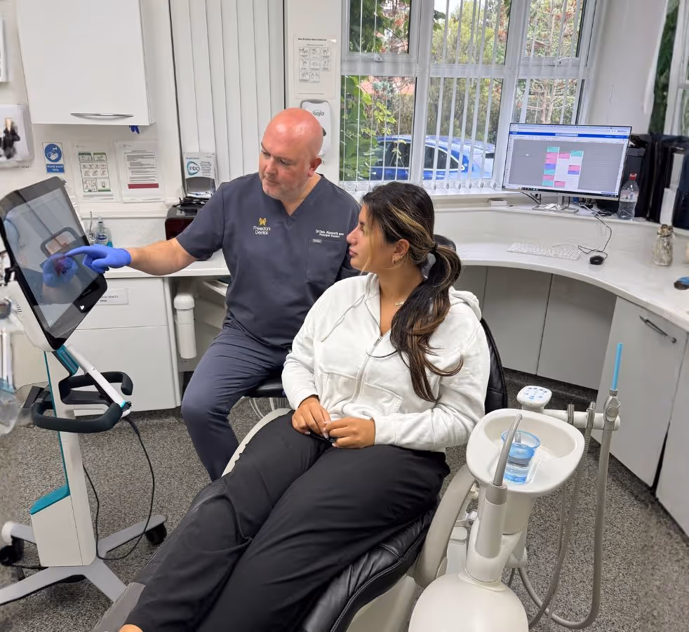 Dentist in navy scrubs showing a patient seated in a dental chair information on a touchscreen monitor in a modern dental office.