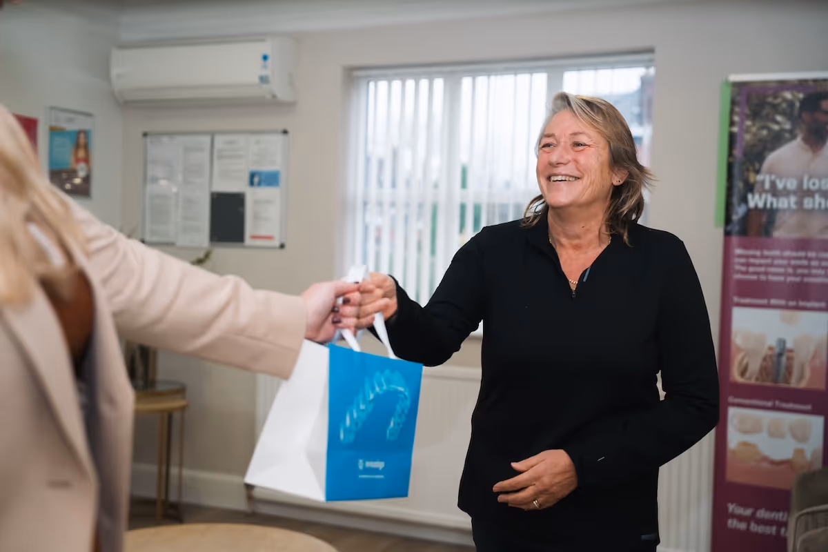Smiling woman in black receiving a dental care bag from another person indoors.