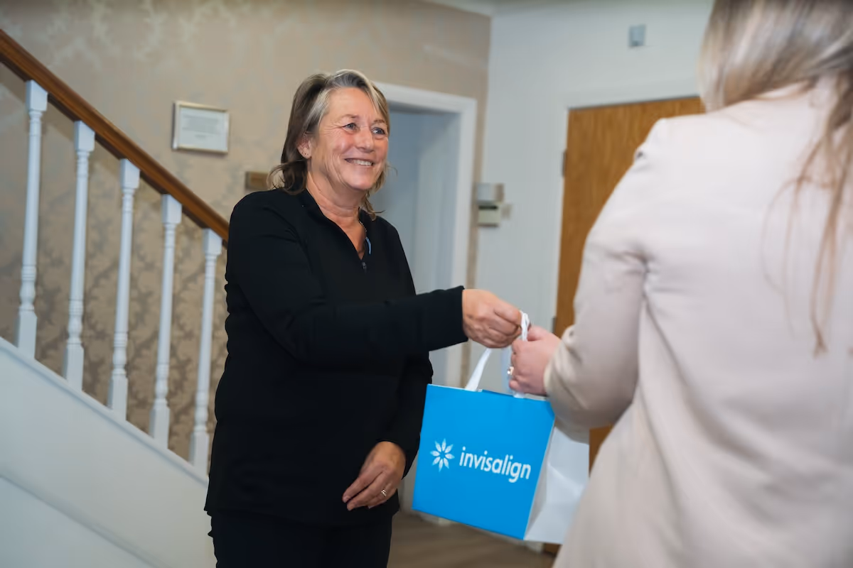 Smiling woman in black handing a blue Invisalign branded bag to another person in a beige jacket indoors near a staircase.